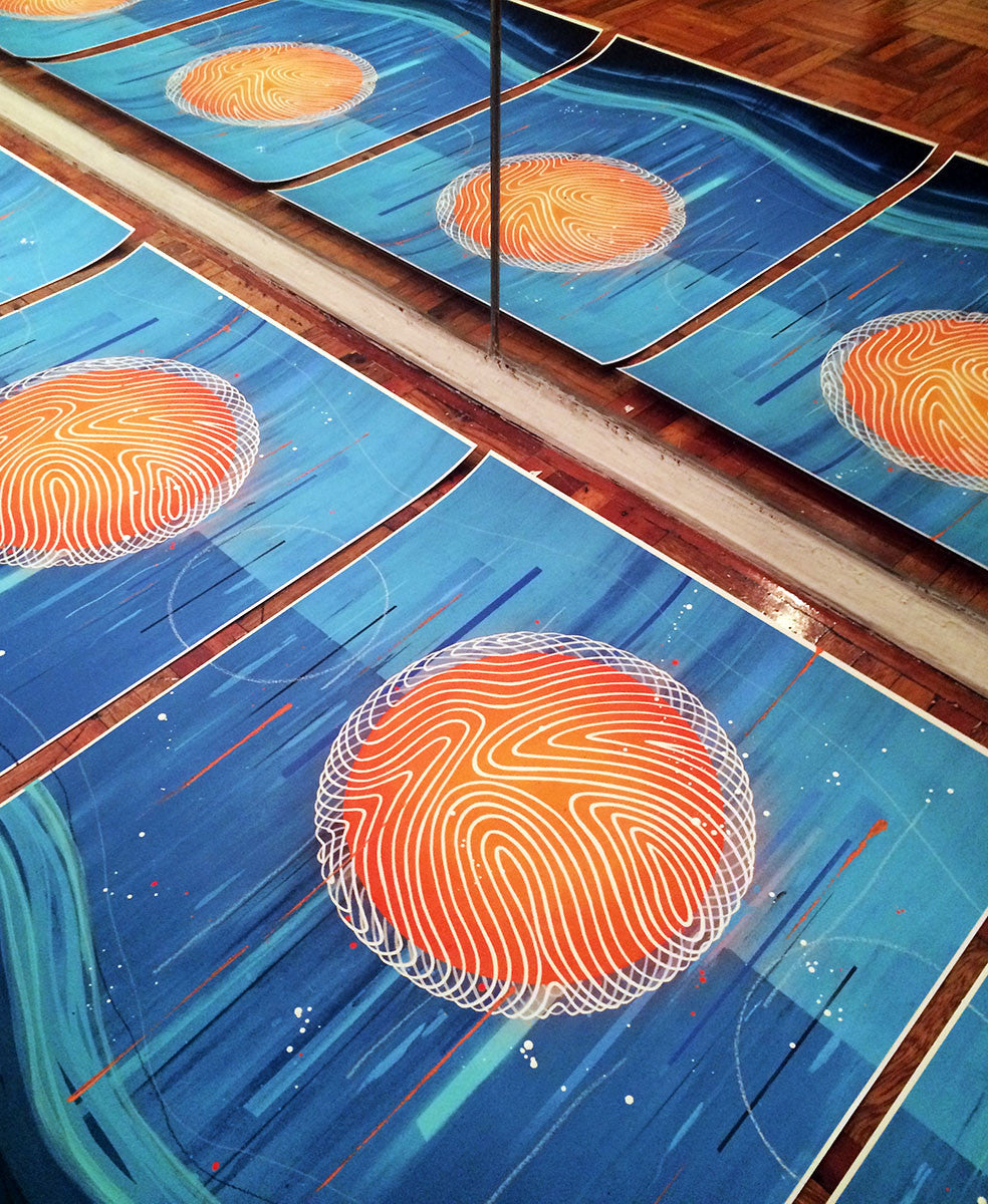 Erik Ottos Change Starts Within art pieces, featuring orange circles and white swirls on a blue background, are displayed on the floor. A mirror reflects these hand-embellished artworks, creating an illusion of more pieces amid streaks of blue and red lines.