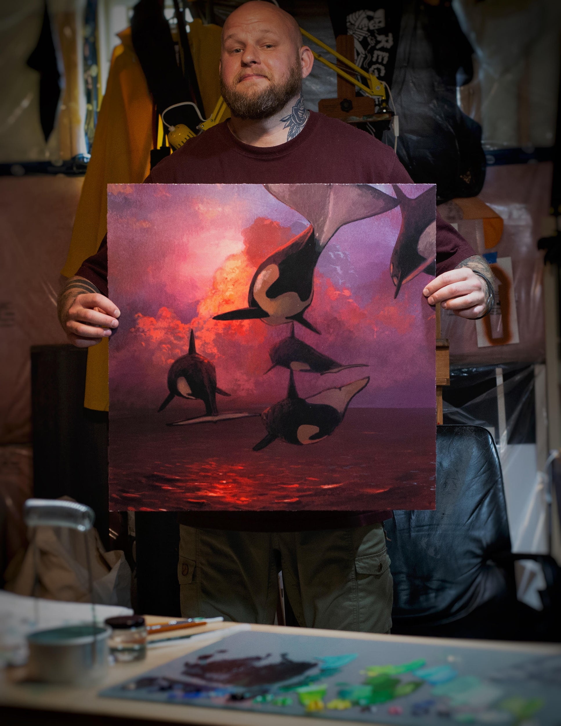 A person holds a hand-embellished edition of Chris Austins Where the Sky Touches the Sea, depicting orcas leaping over a sunset sea. The scene behind them is cluttered with art supplies and canvas, as warm lighting highlights the artworks vibrant colors.
