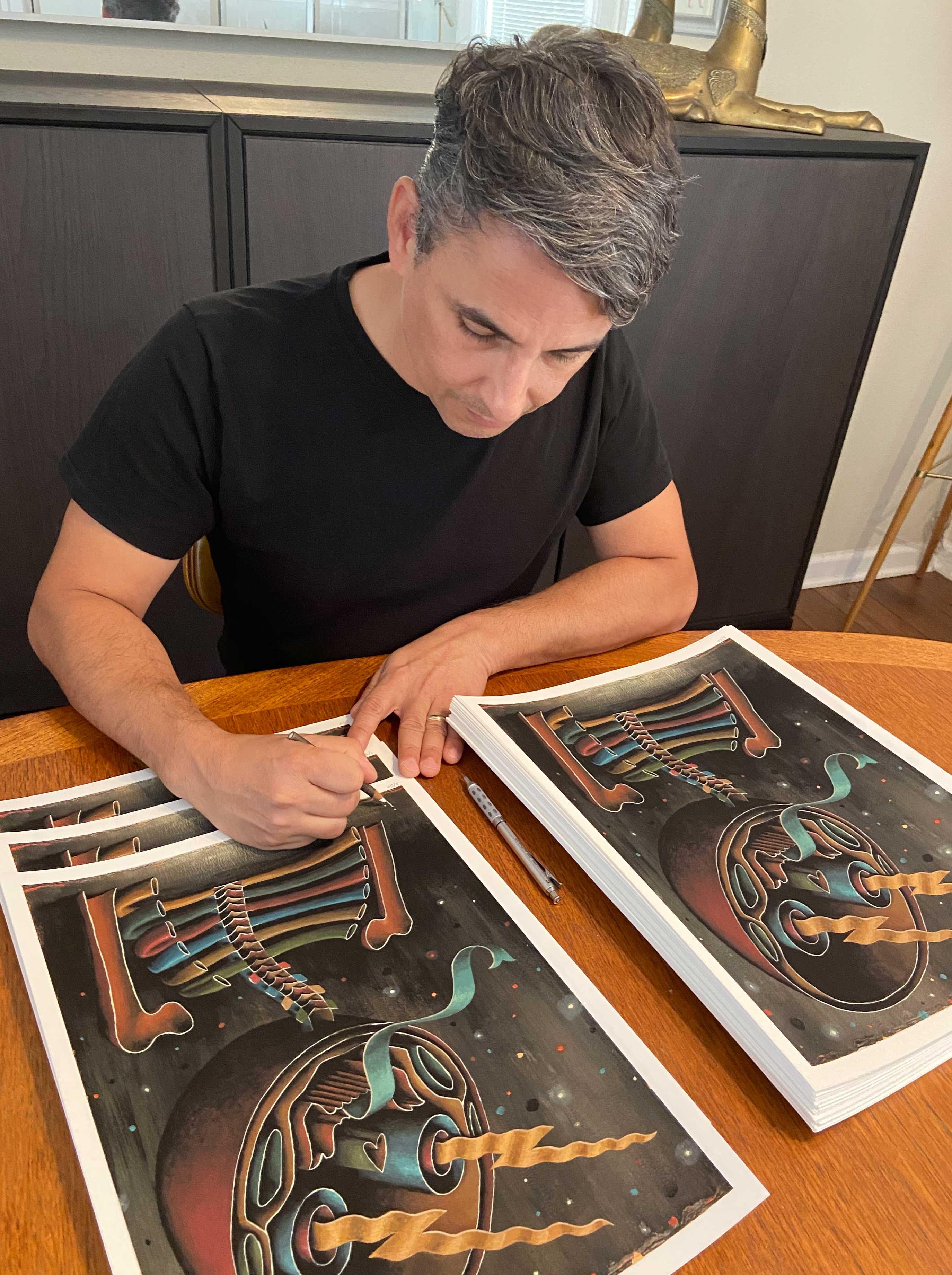 A person with gray hair sits at a wooden table, signing a stack of Bisected archival pigment prints by Jason Limon. Engrossed in their work, theyre dressed in a black T-shirt and holding a pen while the surreal designs with vibrant colors and abstract shapes captivate them.