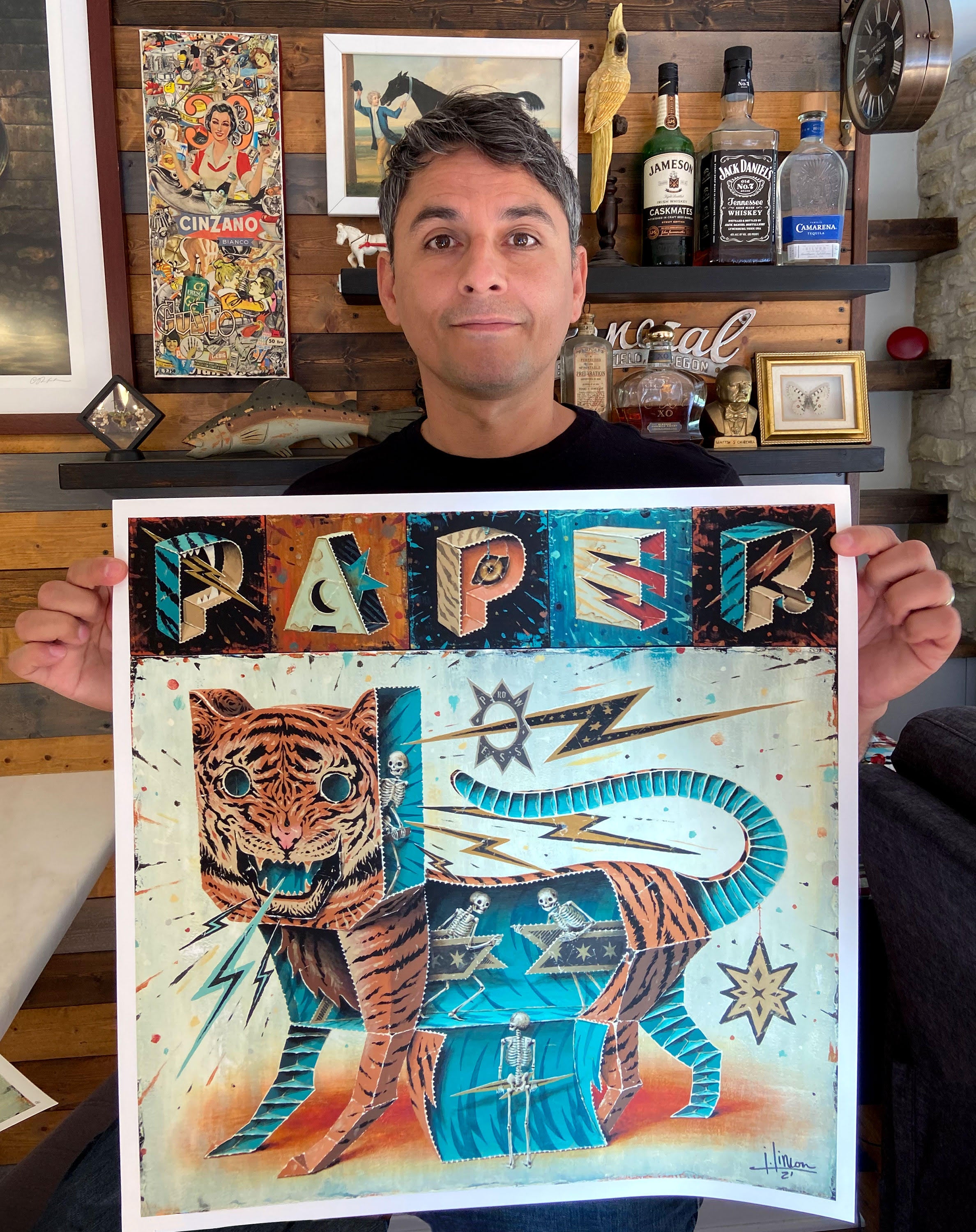 A person holds Prowess, a vibrant archival pigment print by Jason Limon. It features the stylized 3D word PAPER above a tiger with a visible skeleton against an eclectic background of wooden walls, framed images, and bottles. The piece is signed and numbered, enhancing its uniqueness.