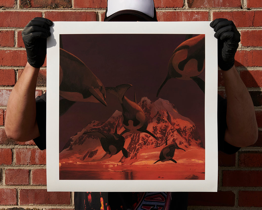 A person wearing gloves and a cap holds In A Silent Way, an archival pigment print by Chris Austin, depicting orcas swimming over a mountainous landscape in reddish hues. The signed, numbered print contrasts with the brick wall backdrop.