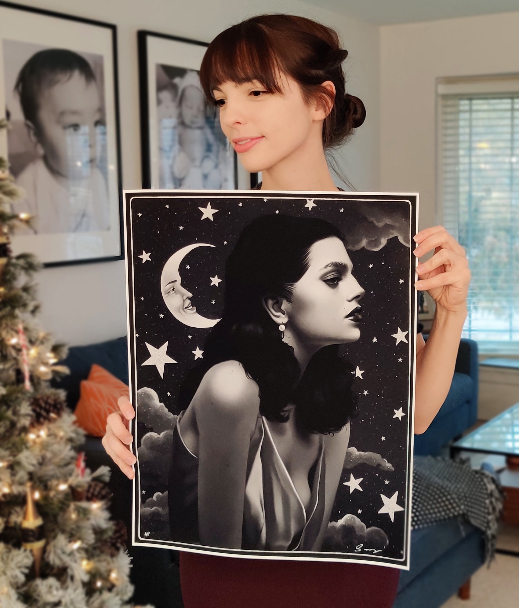 A person holding a signed and numbered archival pigment print titled Worlds A Stage by Sarah Joncas, featuring a black and white profile of a woman with stars and a moon. The background includes a decorated Christmas tree and framed photos on the wall.