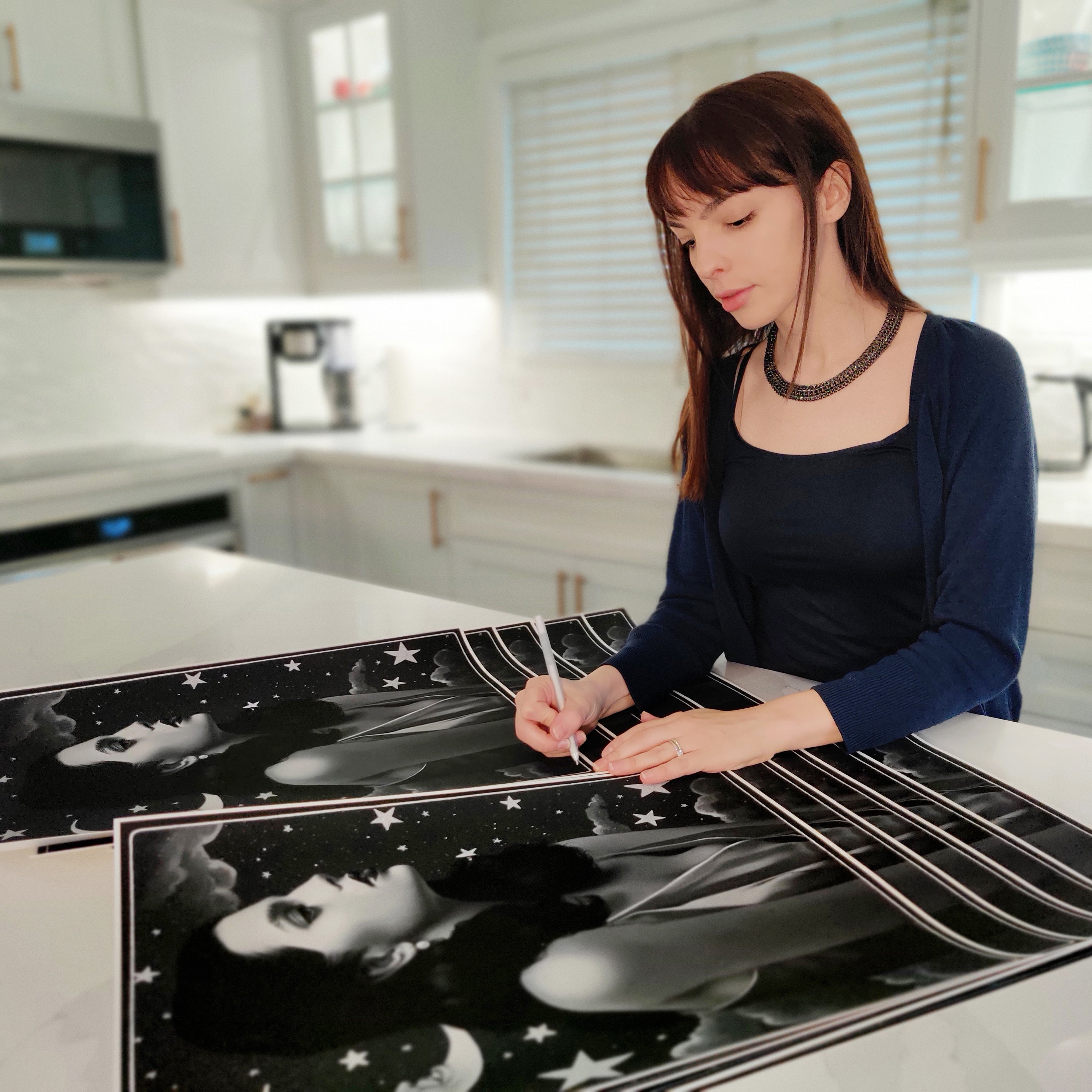 In a modern kitchen featuring white cabinets and stainless steel appliances, Sarah Joncas signs and numbers Worlds A Stage archival pigment prints. Dressed in a black top and blue cardigan, her attention to detail brings the starry night scene with a persons profile to life.