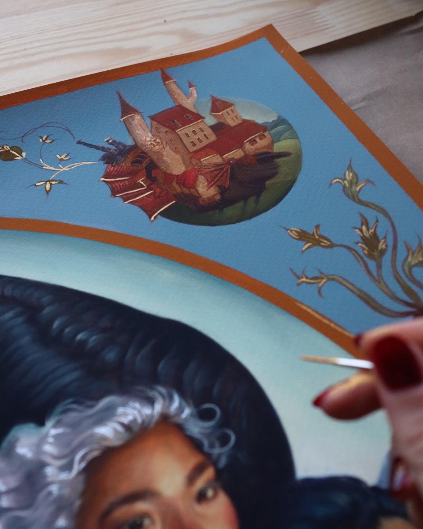 A hand holding a paintbrush meticulously embellishes A Hearts A Heavy Burden by Ruth Speer. The artwork, an archival pigment print, showcases a person in a dark hat and curly hair alongside a fantasy castle scene, creating an exquisite hand-embellished edition.