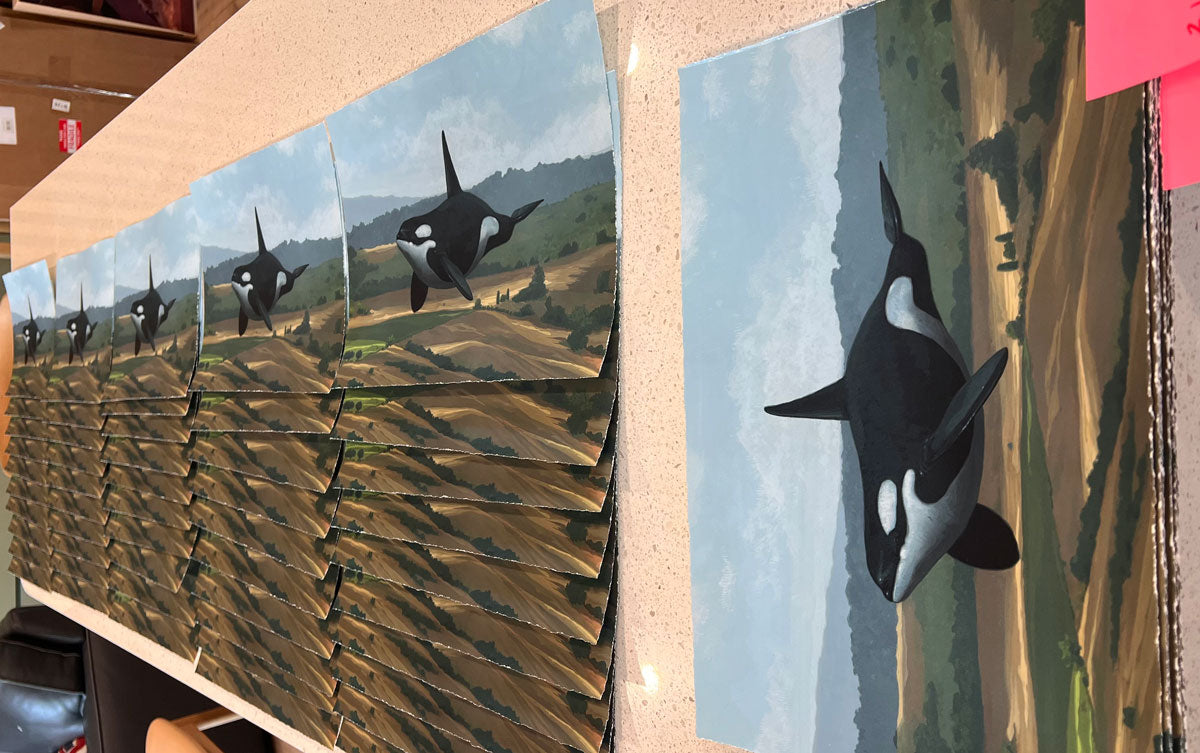 Get On With It" by Chris Austin is an archival pigment print featuring an orca swimming above a landscape with a blue sky and rolling hills in the background. Multiple identical prints are stacked on a countertop.