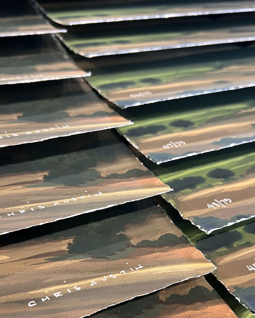 Several "Get On With It" art prints by Chris Austin, featuring green and brown landscapes with torn edges, are stacked in rows. Each archival pigment print is signed and numbered in white.