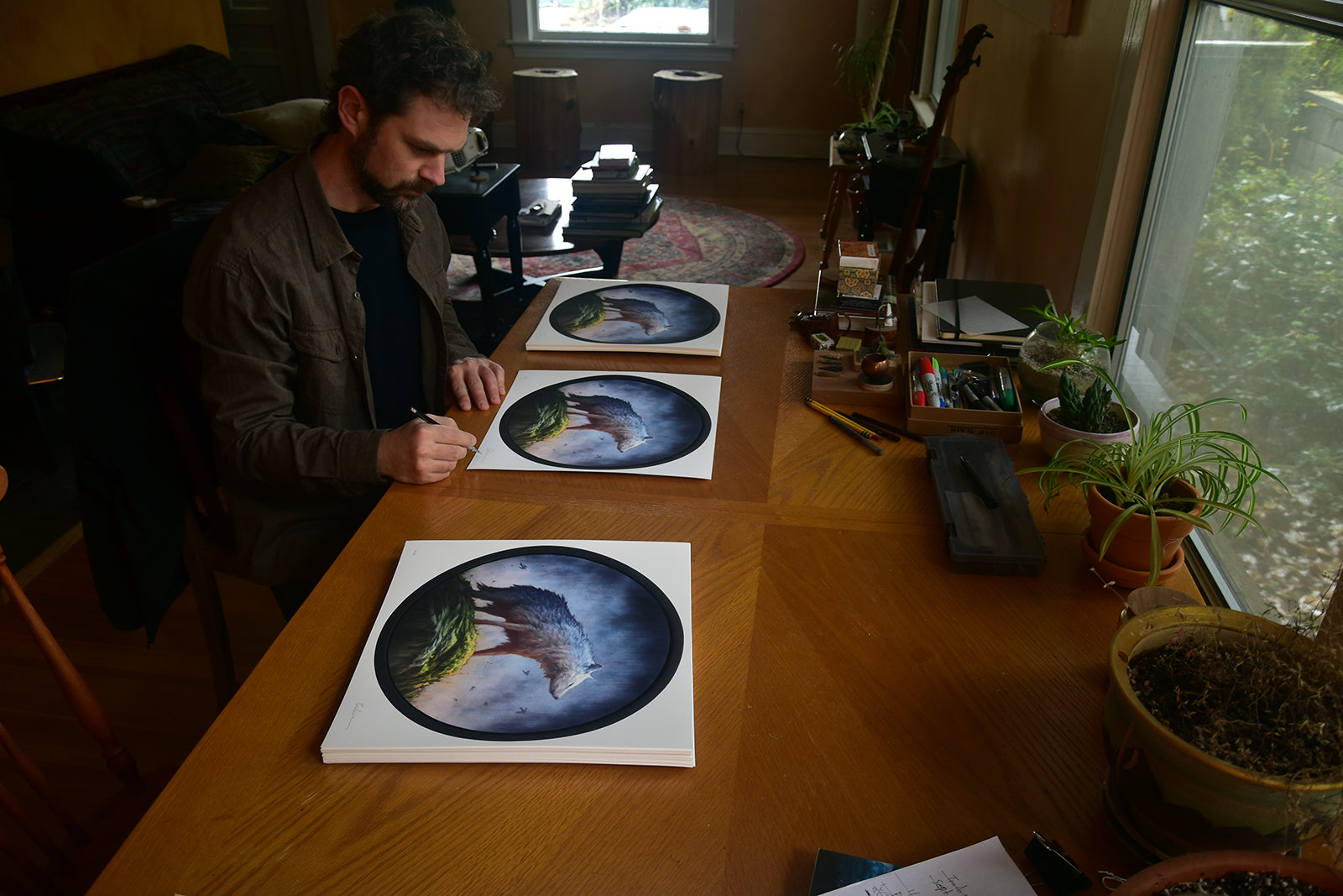 In a sunlit room filled with plants and art supplies, someone sits at a wooden table to sign and number Brin Levinsons archival pigment print titled Winter, which features a circular design of a wolf and foliage, while natural light streams through the large window.