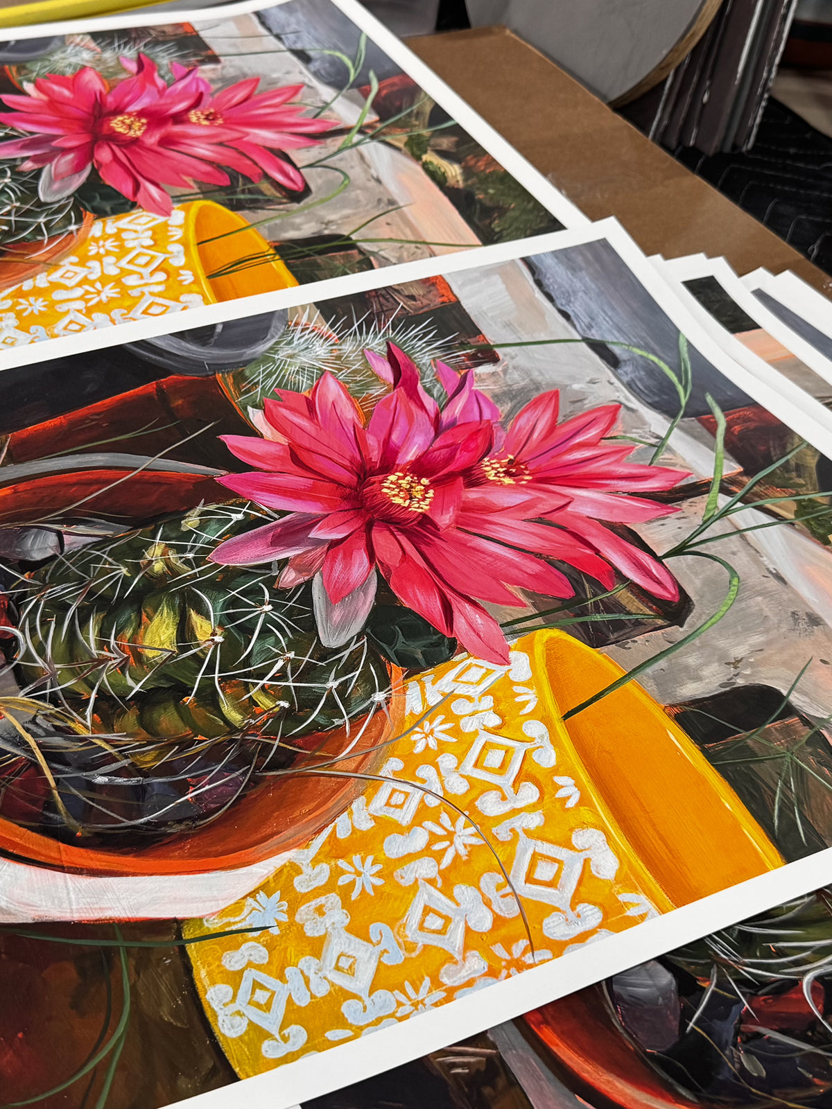 Garden“ by Frank Gonzales features a vibrant cactus with pink flowers in an orange pot on a patterned cloth. Archival pigment prints are shown stacked on a table.