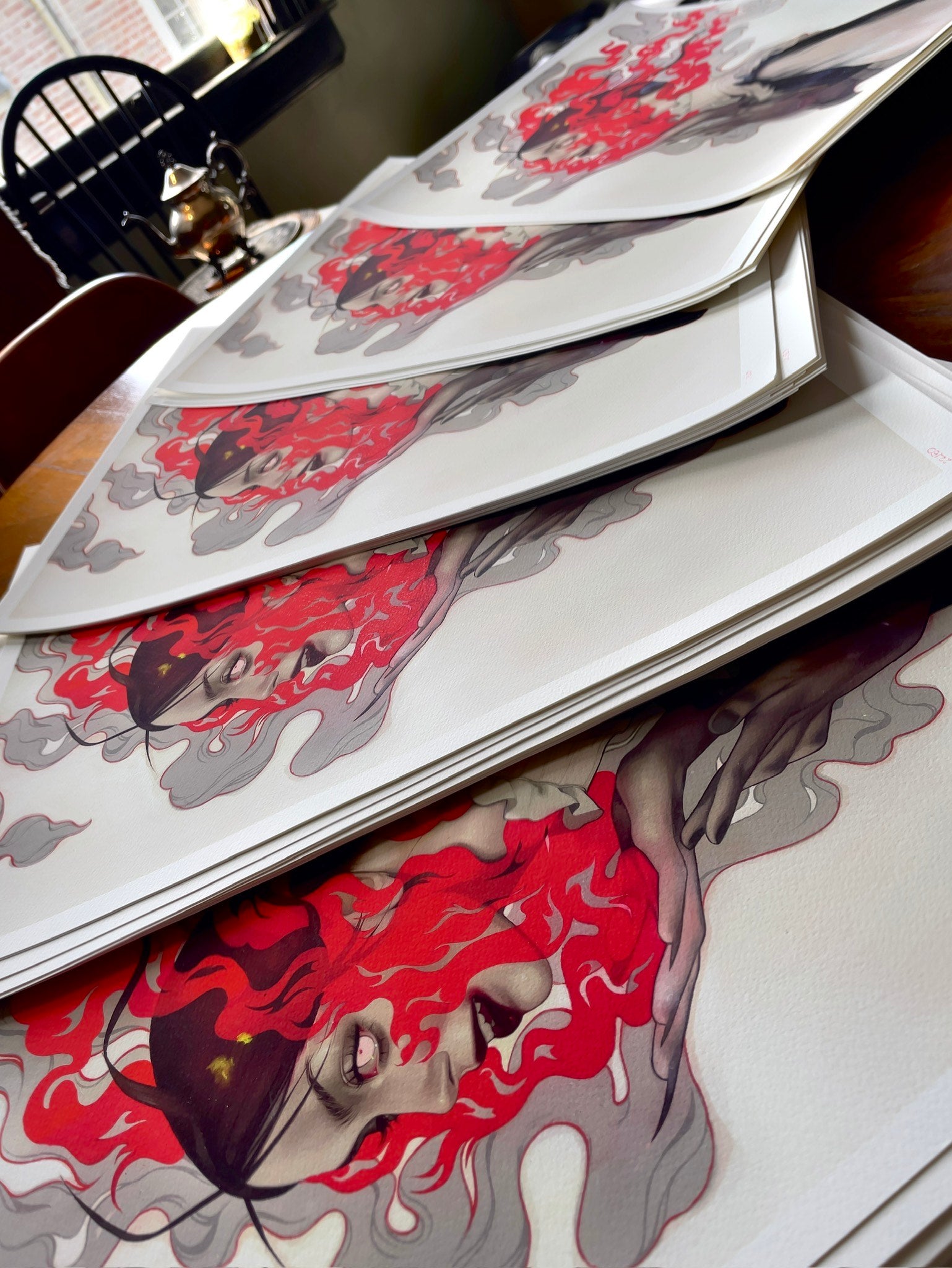 Stacks of Smile for Me, signed and numbered archival pigment prints by Kelsey Beckett, showcase a woman with fiery red hair framed by swirling gray and red smoke-like patterns for a dramatic, surreal effect. These works are displayed on a wooden surface.