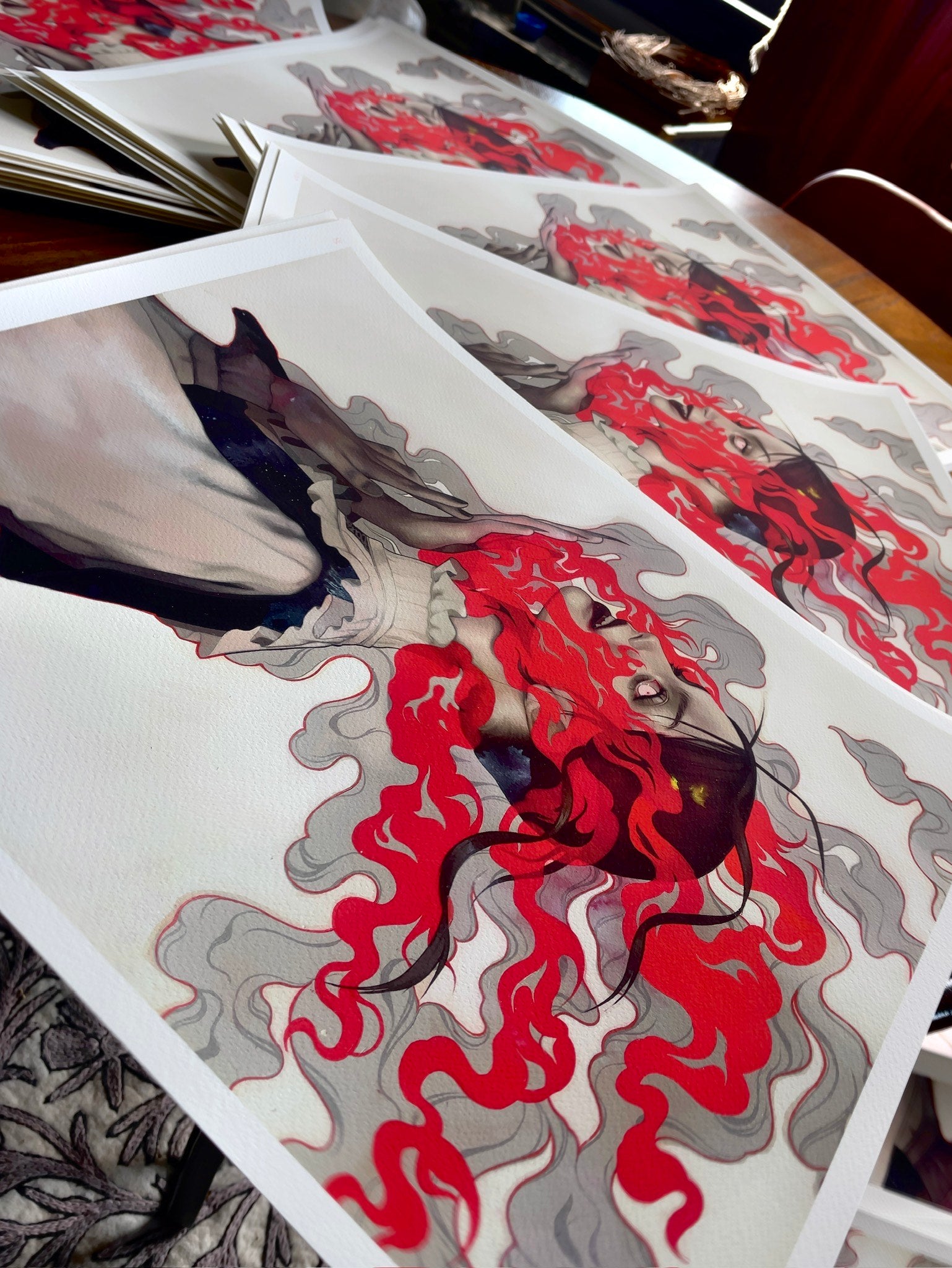 Smile for Me by Kelsey Beckett features a profile of a woman with flowing red hair merging into grey smoke. These archival pigment prints, set against warm lighting, are signed and numbered, with multiple copies displayed on the table.