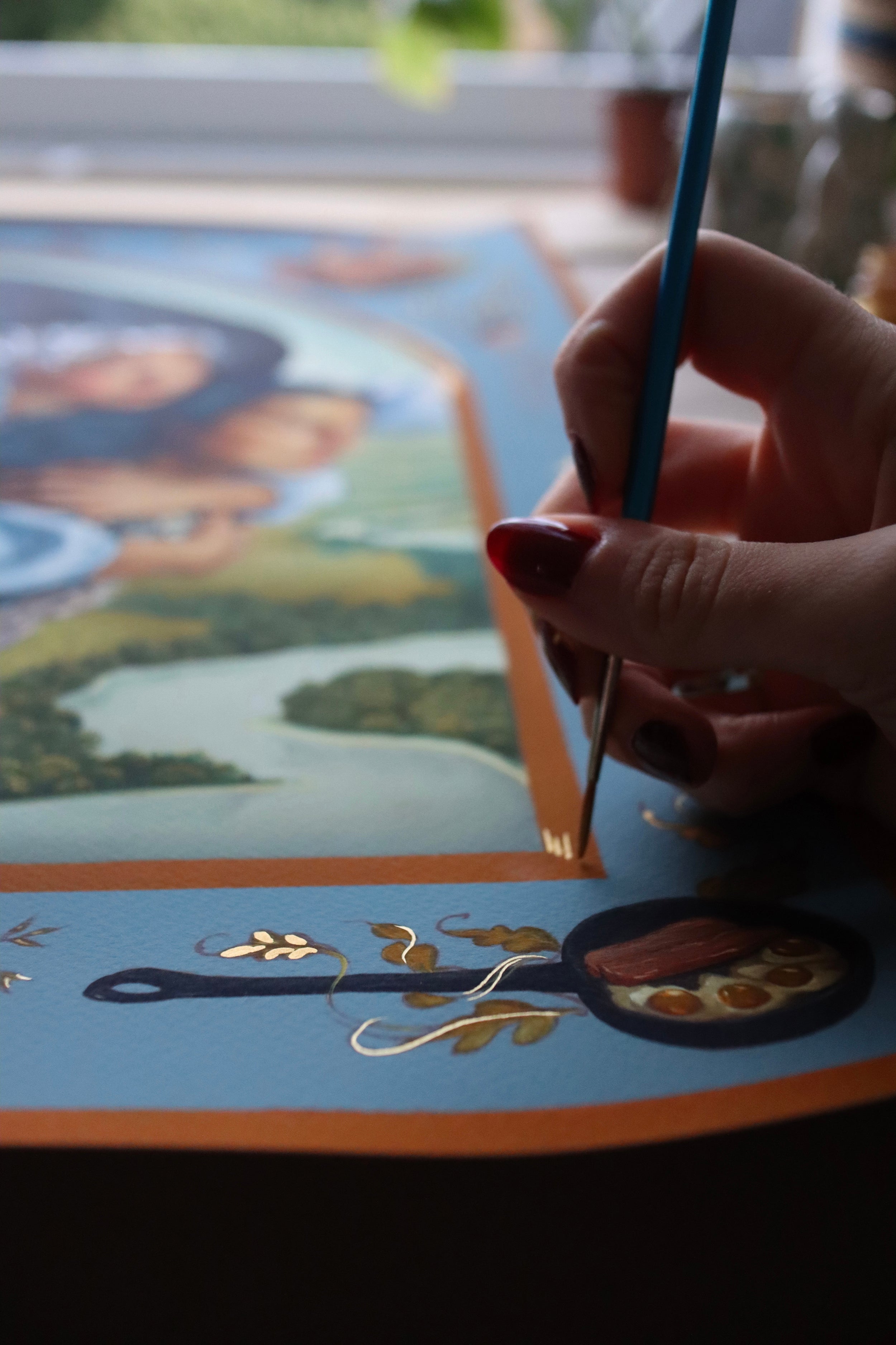 A hand with painted nails, likely Ruth Speers, delicately uses a fine brush to add golden details to an ornate blue artwork titled A Hearts A Heavy Burden, part of her Hand Embellished Edition featuring archival pigment print landscapes and objects.