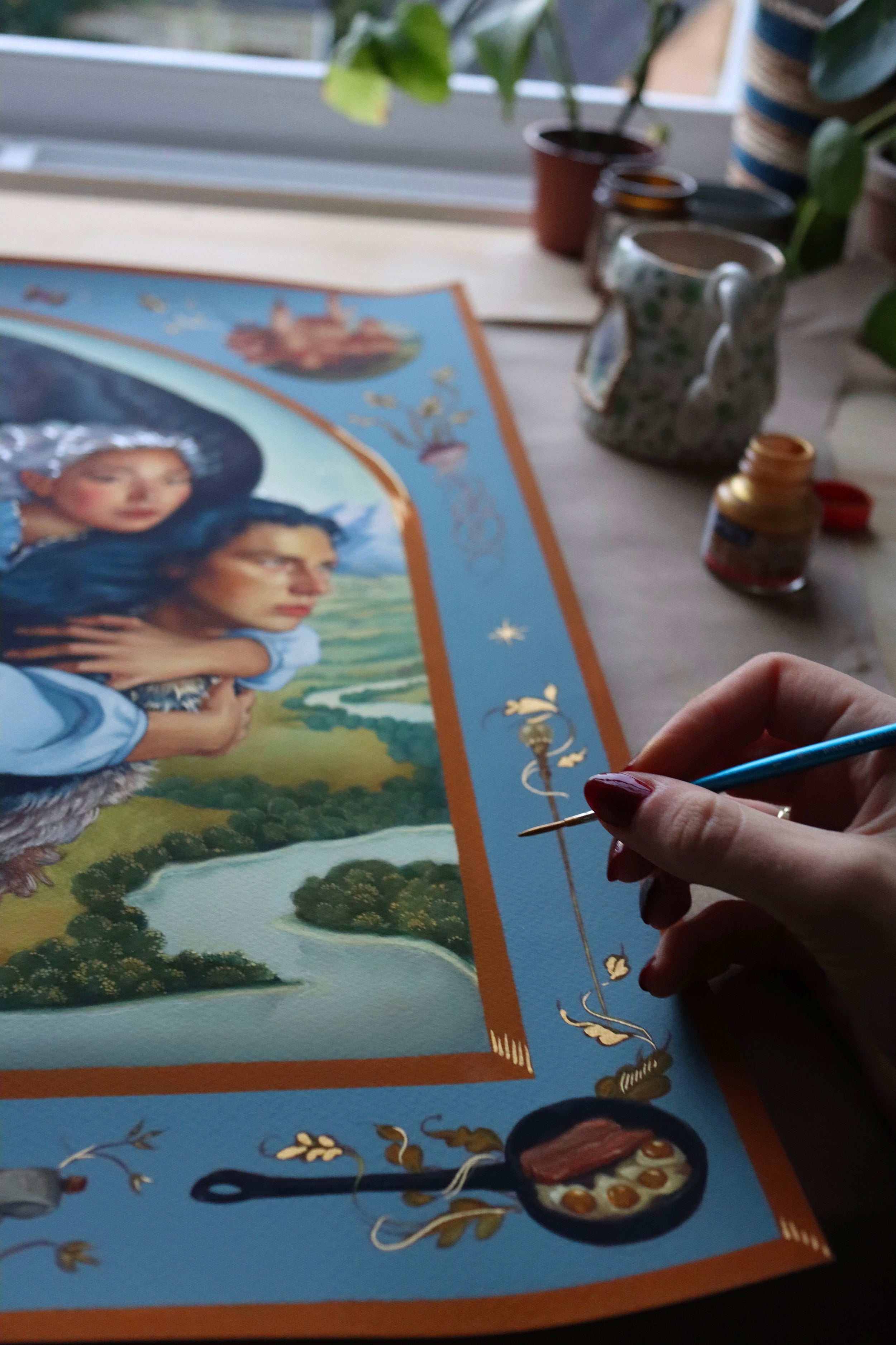 A hand with painted nails delicately embellishes Ruth Speers vibrant A Hearts A Heavy Burden, featuring women, a river landscape, and gold designs. Art supplies are scattered around the table amidst potted plants in this exquisite archival pigment print.