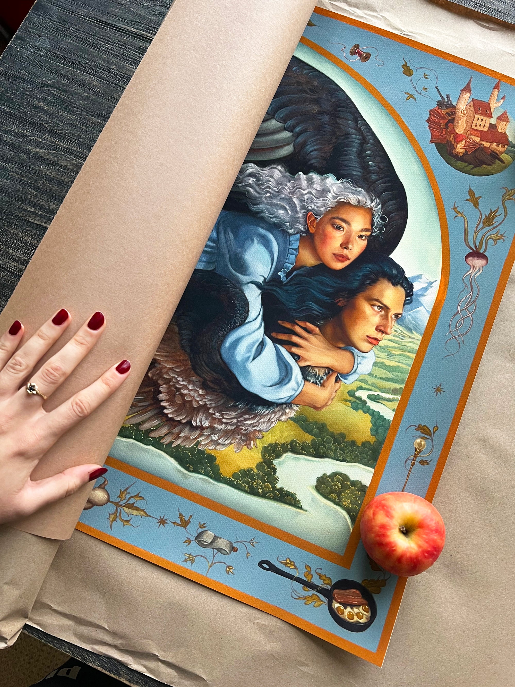 A hand with red nail polish holds a rolled-out Ruth Speer archival pigment print titled A Hearts A Heavy Burden. The artwork shows two winged people embracing while flying over a landscape, bordered by decorative elements and a castle. An apple rests on this hand-embellished edition.