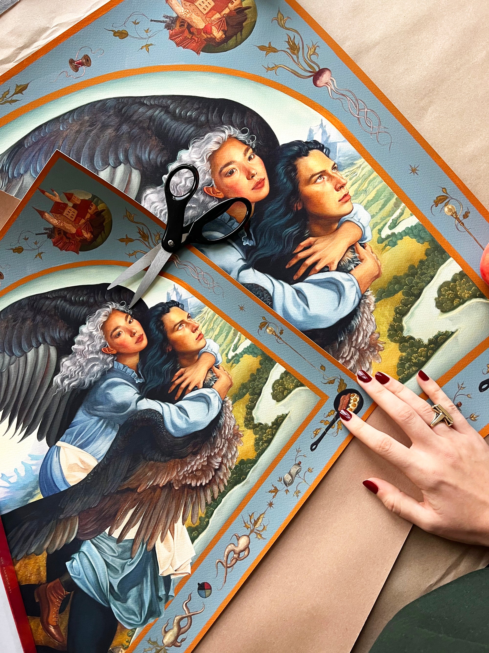 A hand holds a partially cut sheet of Ruth Speers A Hearts A Heavy Burden, showing two winged figures embracing, one with white hair and the other dark. Nearby scissors rest on the Archival Pigment Print, featuring celestial designs in this Hand Embellished Edition.
