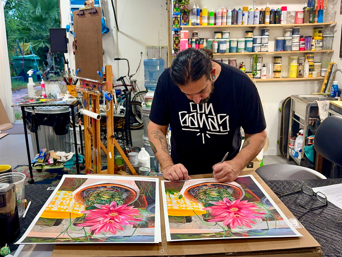 Frank Gonzales signs a vibrant “Garden” floral print in his art studio, surrounded by colorful paint bottles, art supplies, and creative tools as he prepares a hand embellished piece.