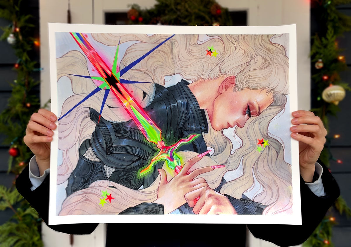 A person holds "Galactic Flare" by Kelsey Beckett—an archival pigment print featuring a blonde armored woman with flowing hair, glowing sword, and bright stars, set among holiday greenery and ornaments.