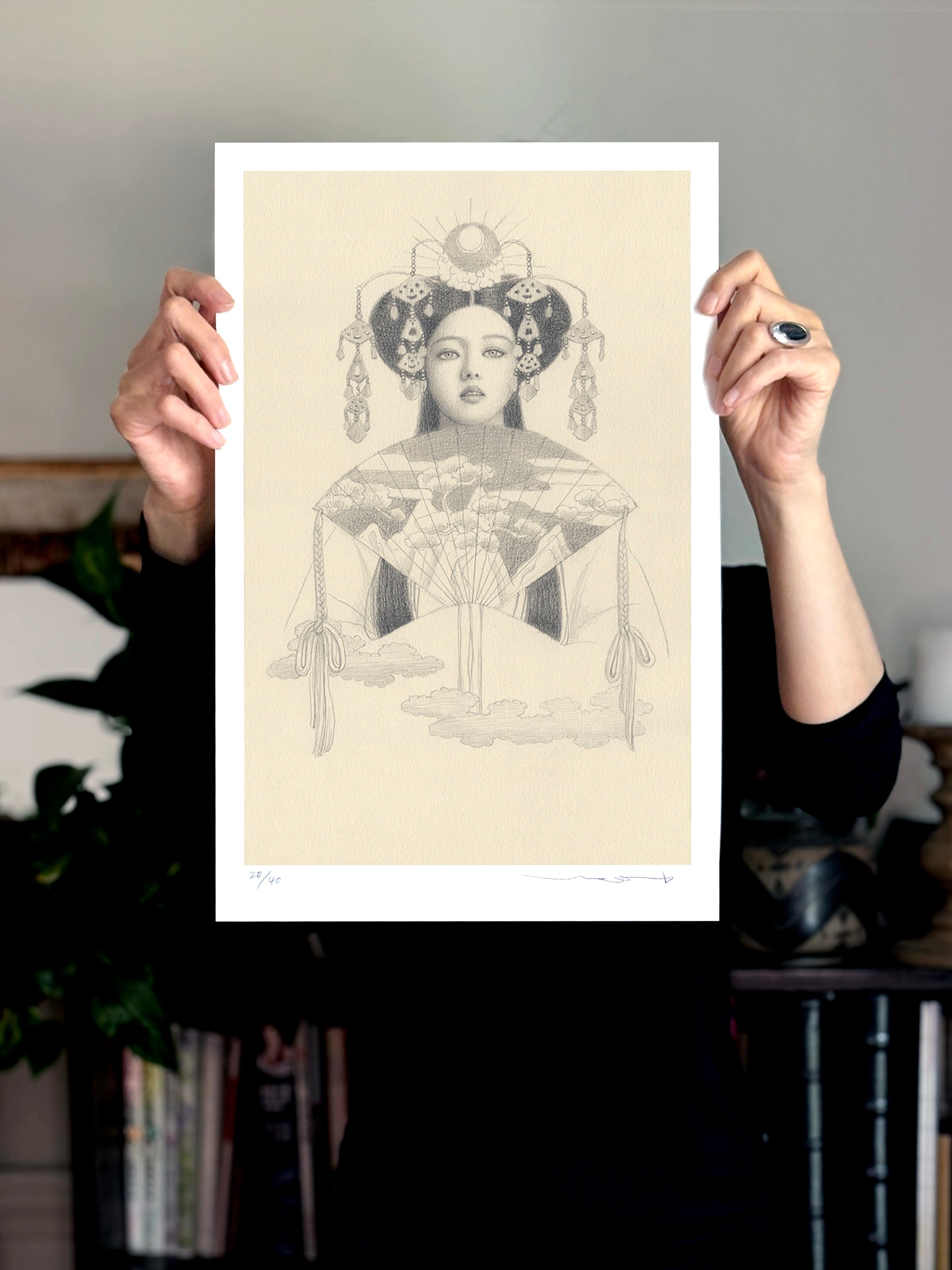 A person in black attire holds up Hemera by Chie Yoshii—a detailed black-and-white drawing on cream paper of a woman with an intricate headdress and fan-like cloud motifs; the holder’s face remains hidden.