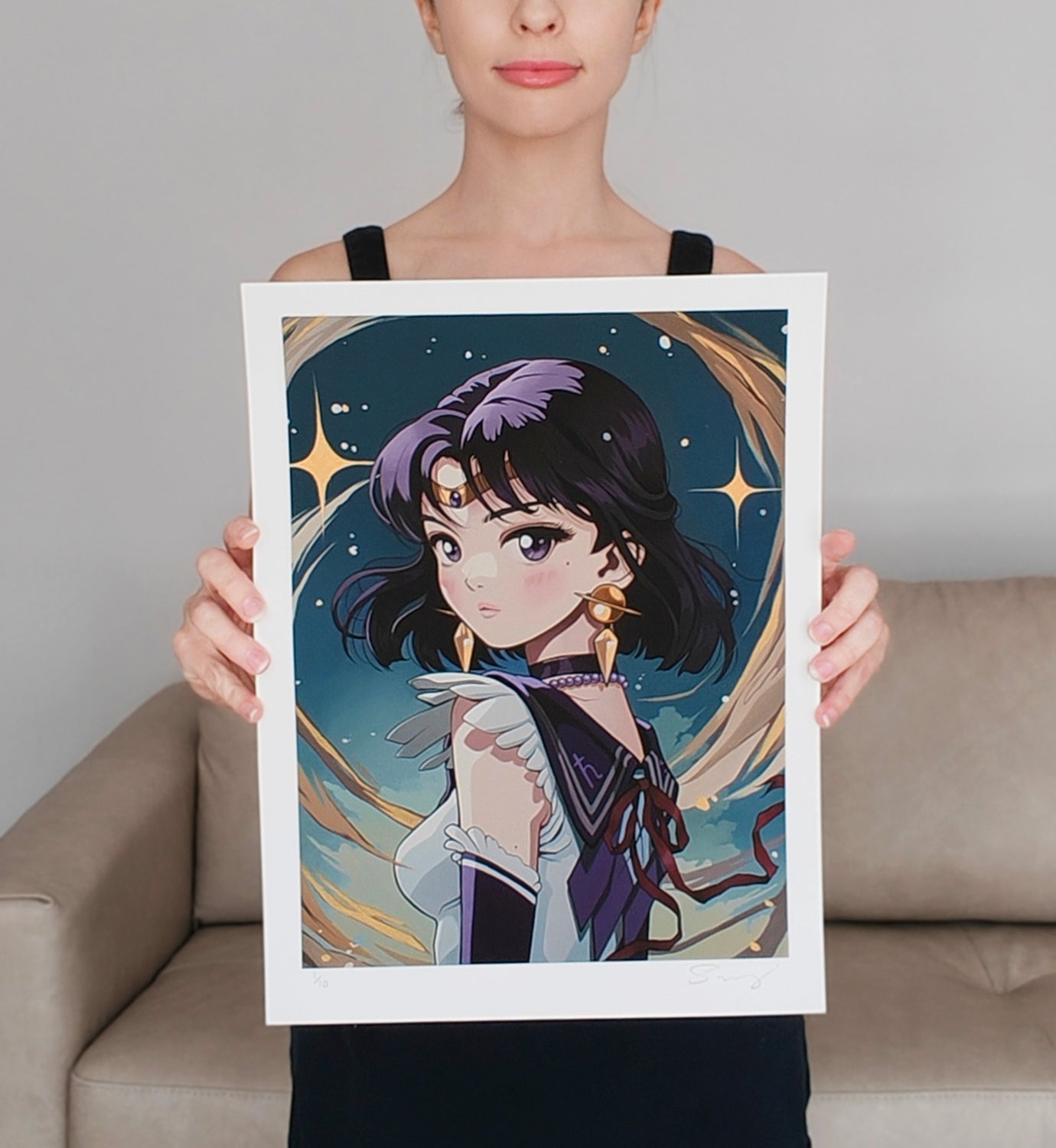 A woman in a black top holds up Sailor Saturn by Sarah Joncas, an anime-style illustration with a cosmic background and gold accents, depicting Sailor Saturn with short dark hair, a headpiece, and earrings.