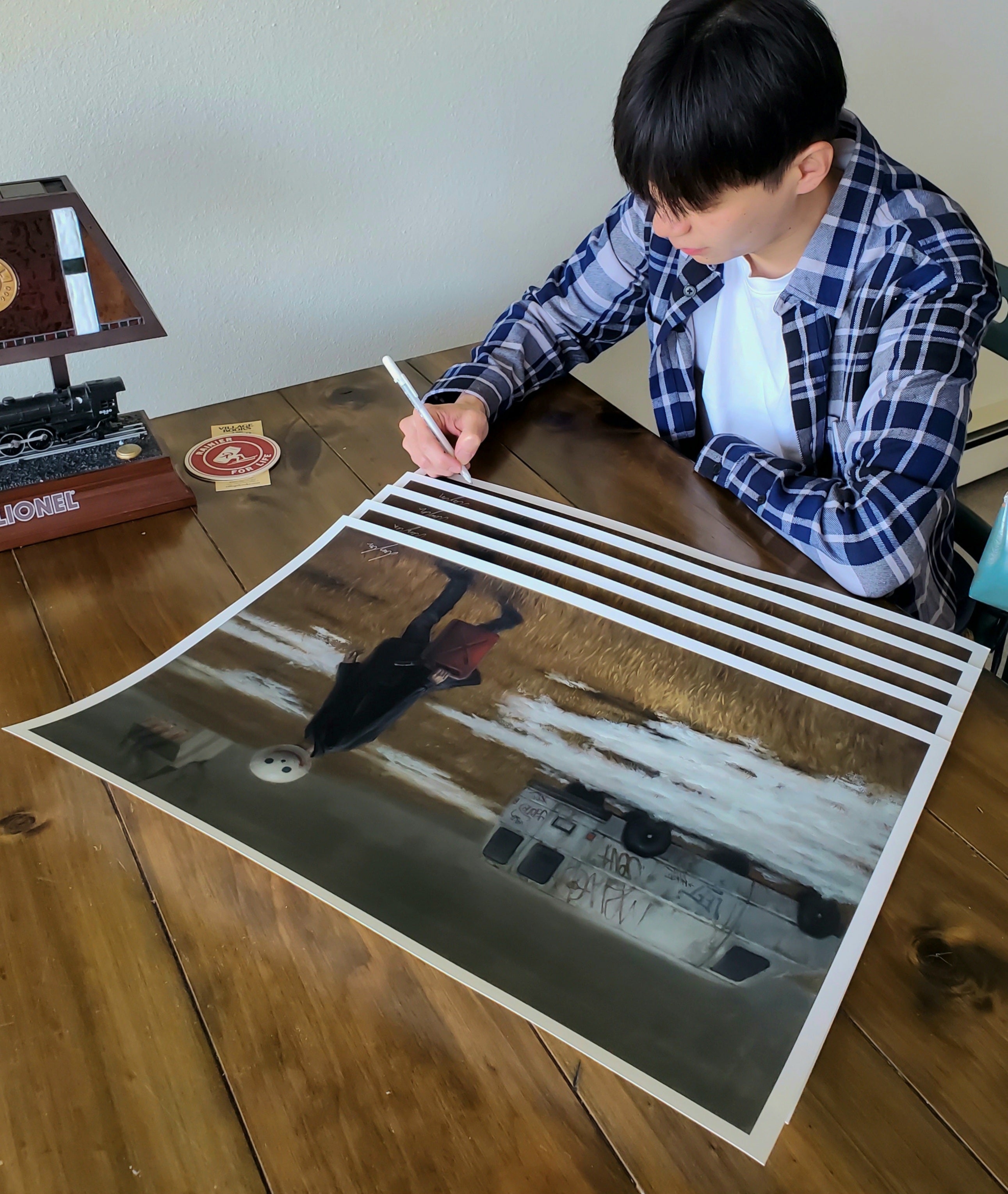 A person in a plaid shirt and white t-shirt signs a limited edition Burning print by Leegan Koo at a wooden table. This artwork showcases a scarecrow and van, with a model train and coaster nearby.
