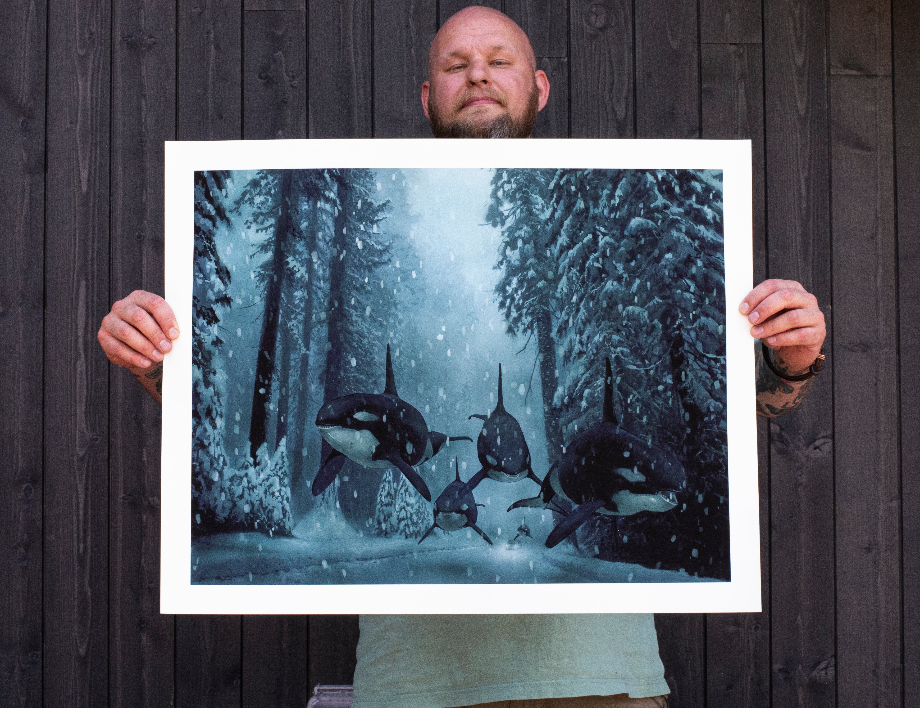 A person holds A Quiet Dream, a signed and numbered Chris Austin artwork, showing orcas swimming through a snowy forest with tall trees and falling snowflakes, captured in vivid detail using archival pigment print.