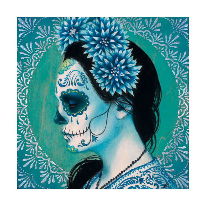 A person with Day of the Dead face paint in blue and green hues is adorned with Blue Dhalia by Sylvia Ji. Blue dahlias decorate their dark hair, complementing a beaded necklace, while a circular teal and white pattern frames the scene, creating an archival art print effect.