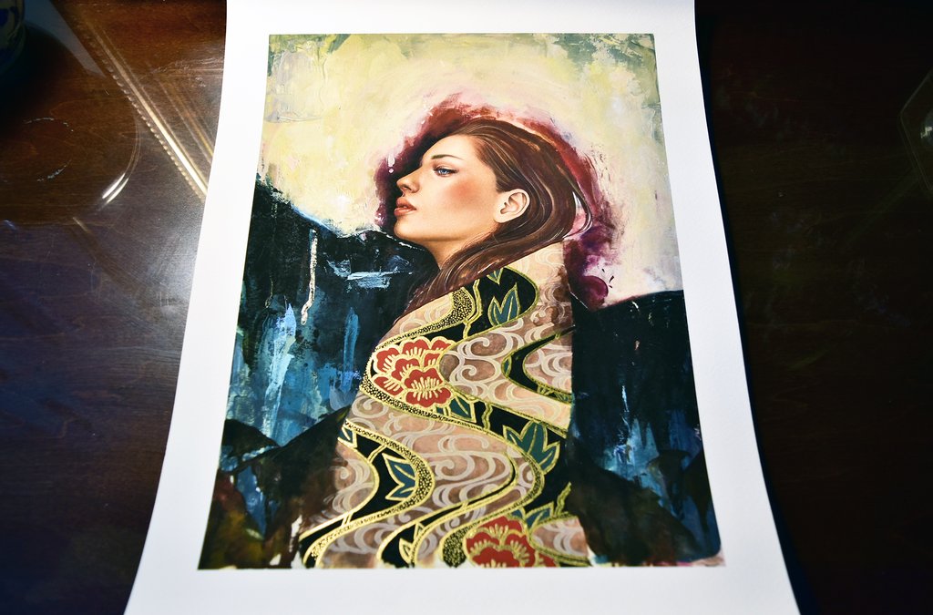 Linger by Sasha Ira features a woman with long hair in side profile donning a cloak with floral motifs against a dramatic backdrop of light and dark hues. Available as a signed and numbered archival pigment print for exclusivity.