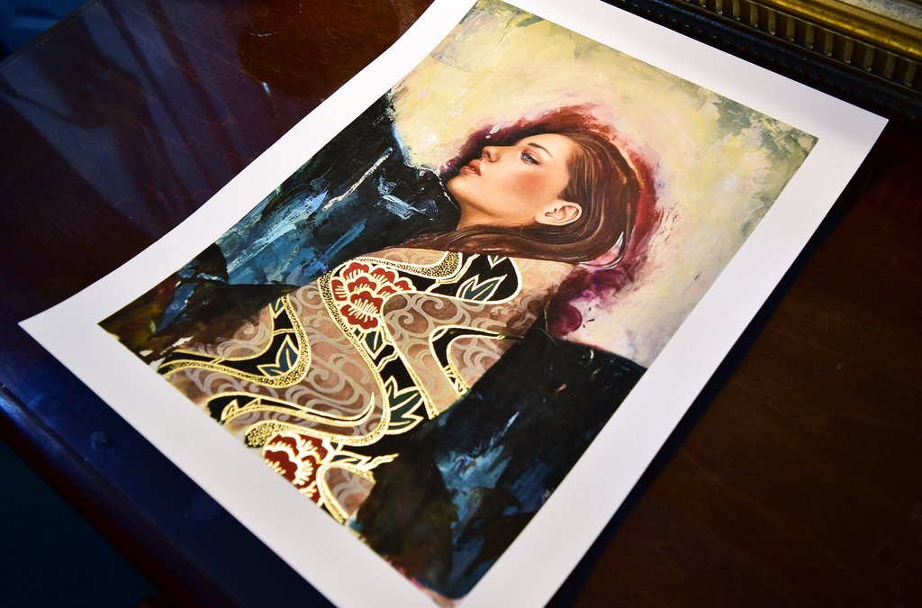 Linger by Sasha Ira is a signed and numbered print depicting a woman in profile with long hair, vibrant colors, and an abstract background. She dons an ornate red and gold patterned garment, resting on a dark wooden surface.