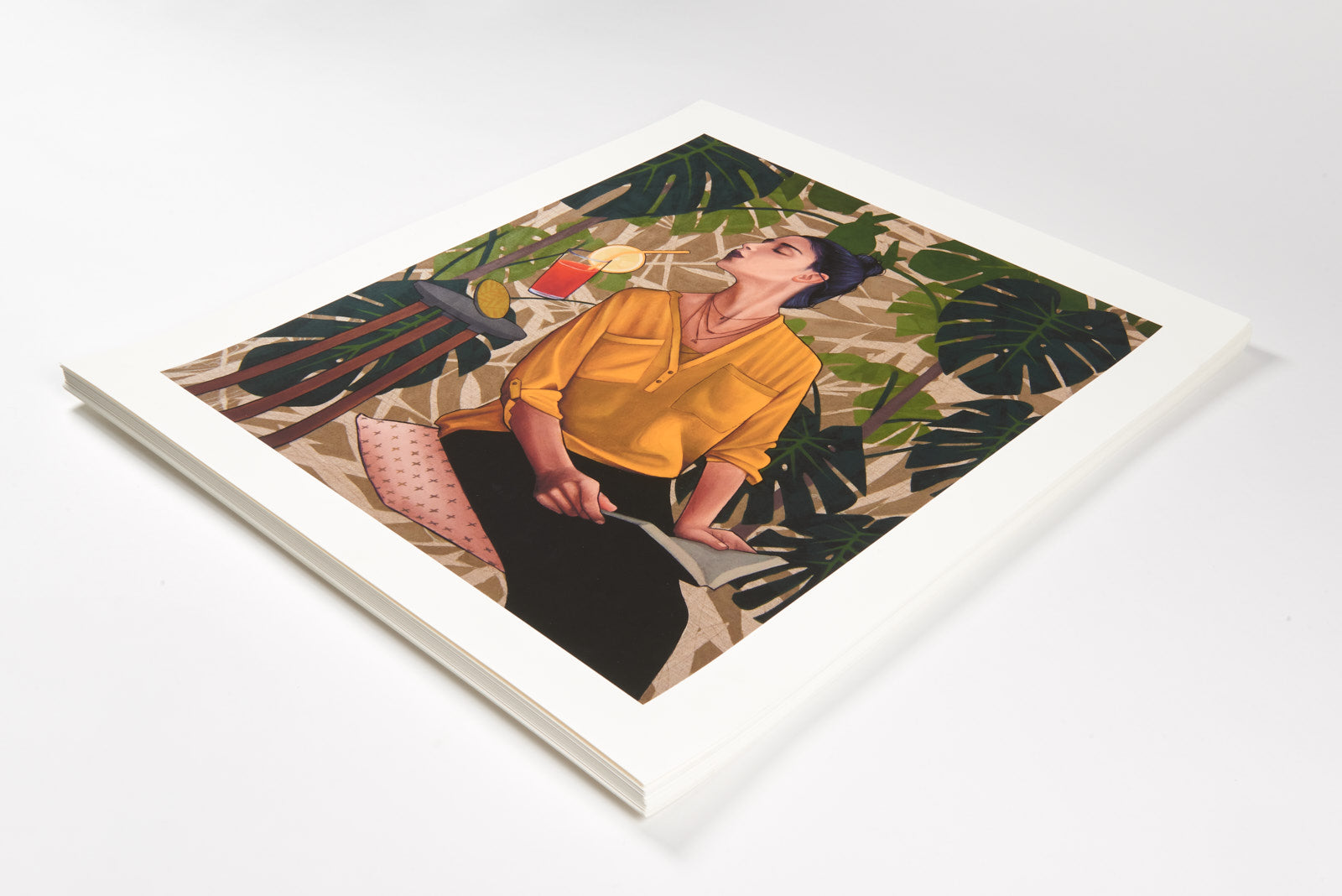 A stack of Artezs hand-numbered, archival pigment prints titled Thirst shows a person in a yellow shirt amidst tropical leaves, holding a fruity drink and gazing upward thoughtfully.
