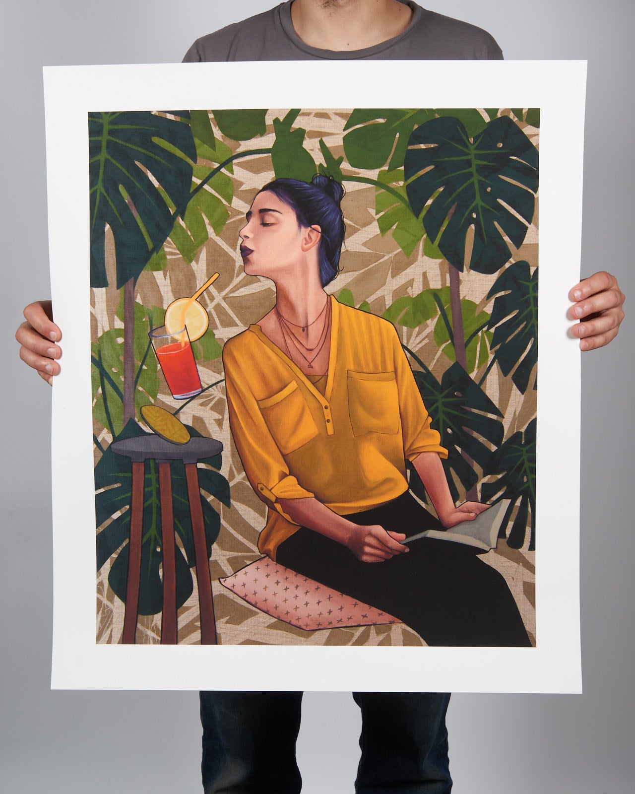 A person holds Artezs Thirst, a large, hand-numbered archival pigment print. It depicts a woman with dark hair in a bun wearing a yellow blouse, sipping a drink with a straw while seated on a stool. The leafy green background enhances the tropical vibe. Includes certificate of authenticity.