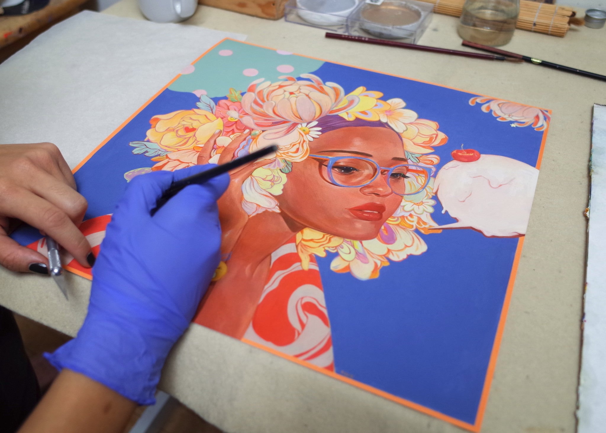 A person with blue gloves paints Sweetness #2 by Helice Wen, showcasing a woman with glasses and intricate floral headwear on a vibrant blue background. The scene on the light-colored table features art supplies like brushes and paints for this hand-embellished masterpiece.
