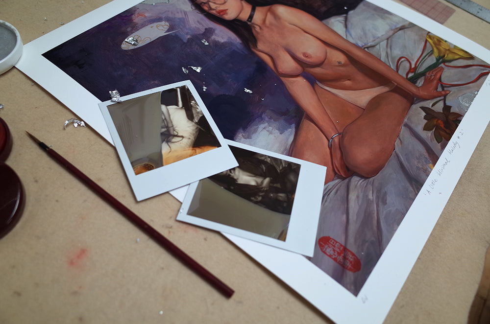 On the table is Little Minimal Melody #2, an archival pigment print by Helice Wen. It features a nude female figure on a bed with a flower, enhanced by dark purple and blue hues. Two Polaroid photos and a thin paintbrush rest on this beautiful masterpiece.