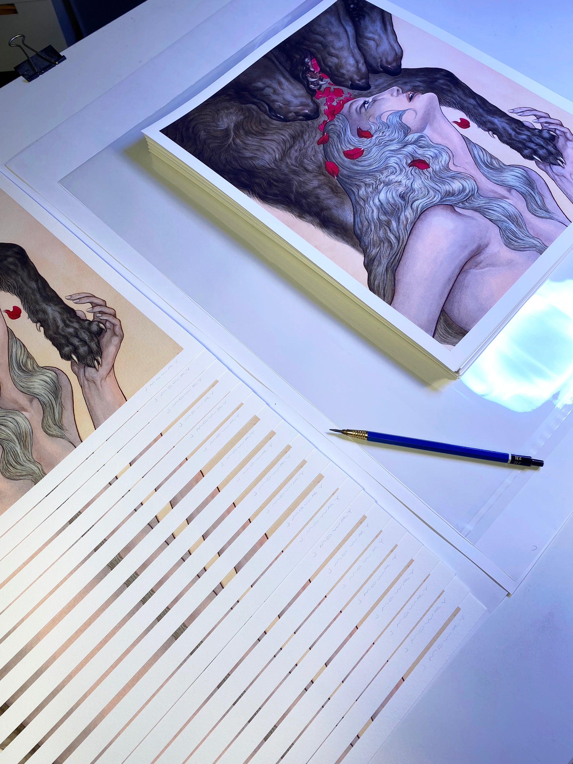 Blessings from the Maw art prints by Jason Mowry show intertwined figures with dark and light hair amid red flowers. A stack of archival pigment prints is beside one being trimmed with a paper cutter, while a pencil rests nearby.