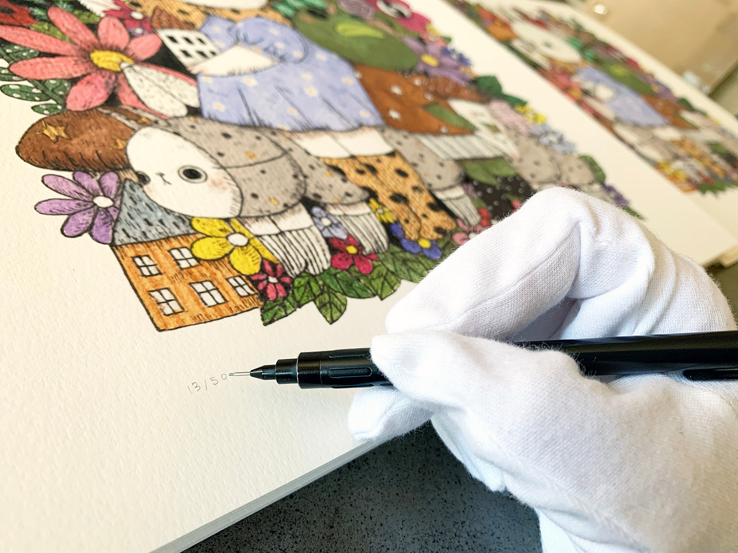 A gloved hand numbers and signs a limited edition archival pigment print of The Gala by jb0xtchi in the corner, featuring colorful, whimsical characters and flowers.