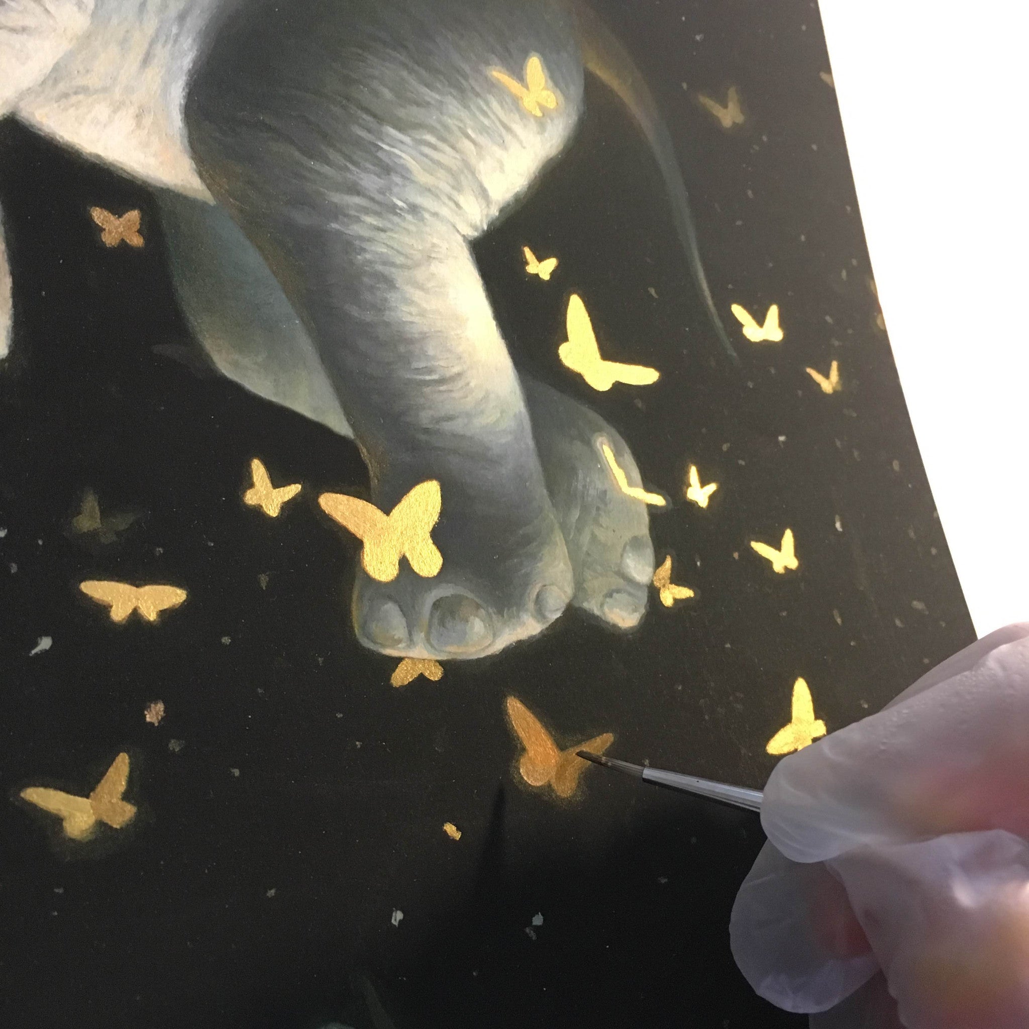 A gloved hand paints a surreal scene, Midnight Ballet by KiSung Koh, where glowing golden butterflies surround an elephants foot against a dark backdrop.
