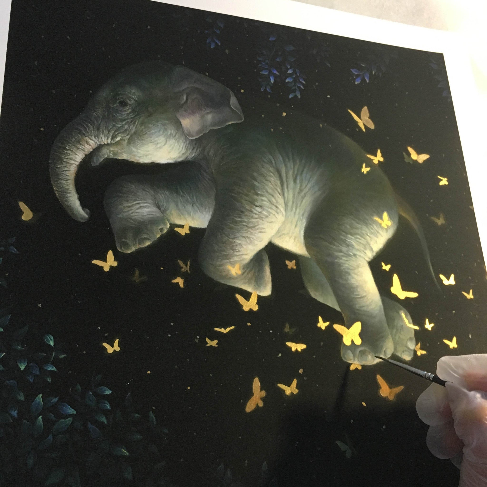 In Midnight Ballet by KiSung Koh, a painting of a floating baby elephant is encircled by glowing butterflies on a dark backdrop. A hand with a paintbrush refines one butterfly, while leaves faintly appear at the edges.