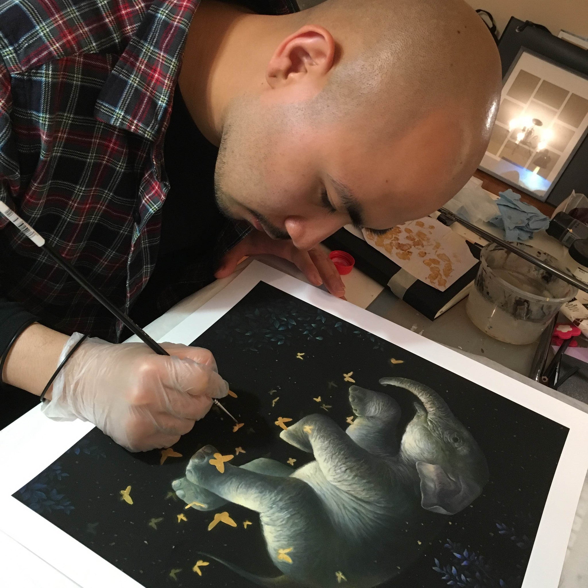 Wearing a plaid shirt, a person paints an elephant with floating butterflies using a fine brush. The artwork echoes KiSung Kohs Midnight Ballet style, with art supplies scattered across the workspace for this hand-embellished piece.