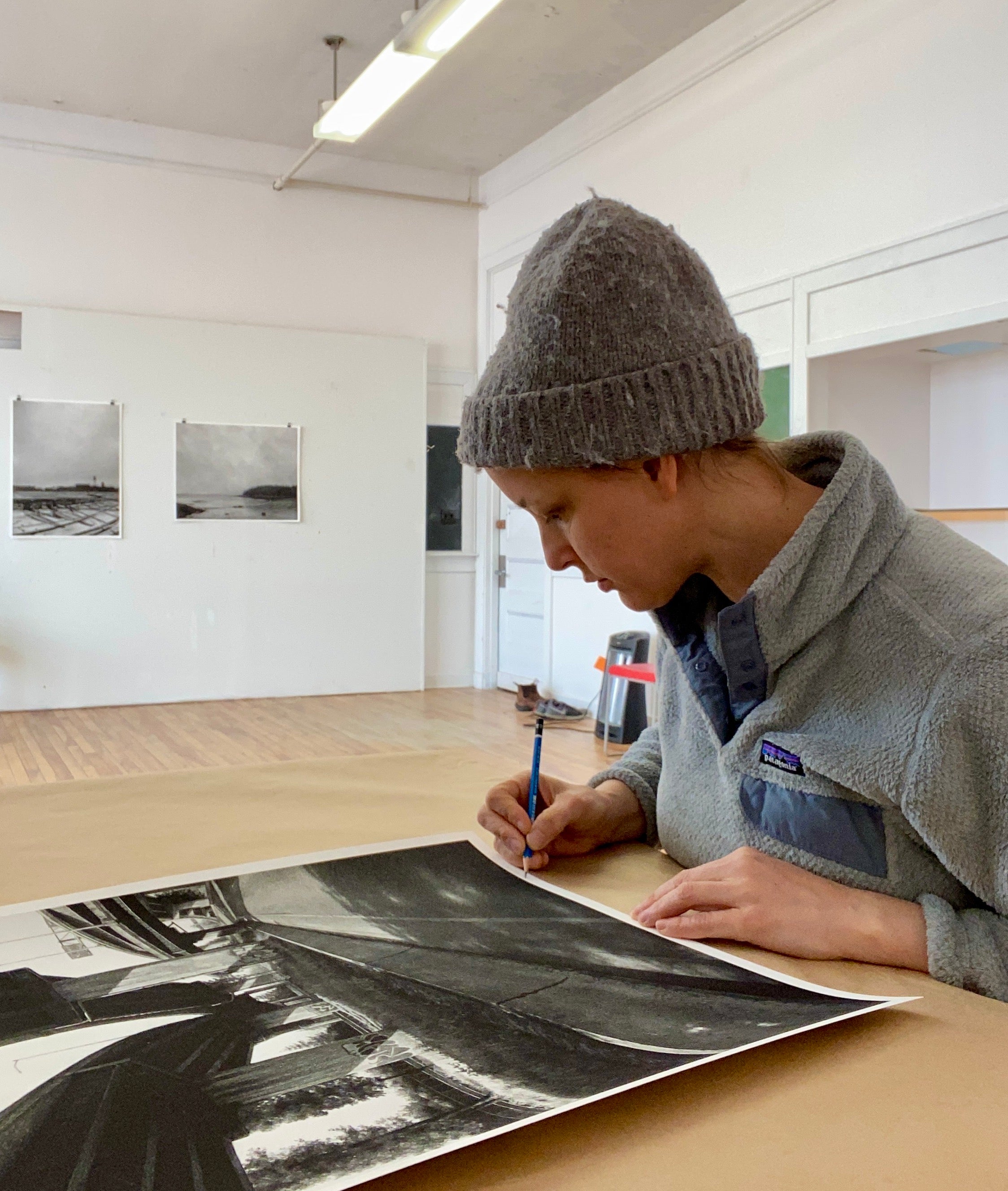 In a spacious, well-lit art studio, a person wearing a wool hat and fleece jacket sketches on a large sheet of paper. Adorning the wall are black-and-white artworks called 5 North by Stephanie Buer, with signed and numbered archival pigment prints nearby.