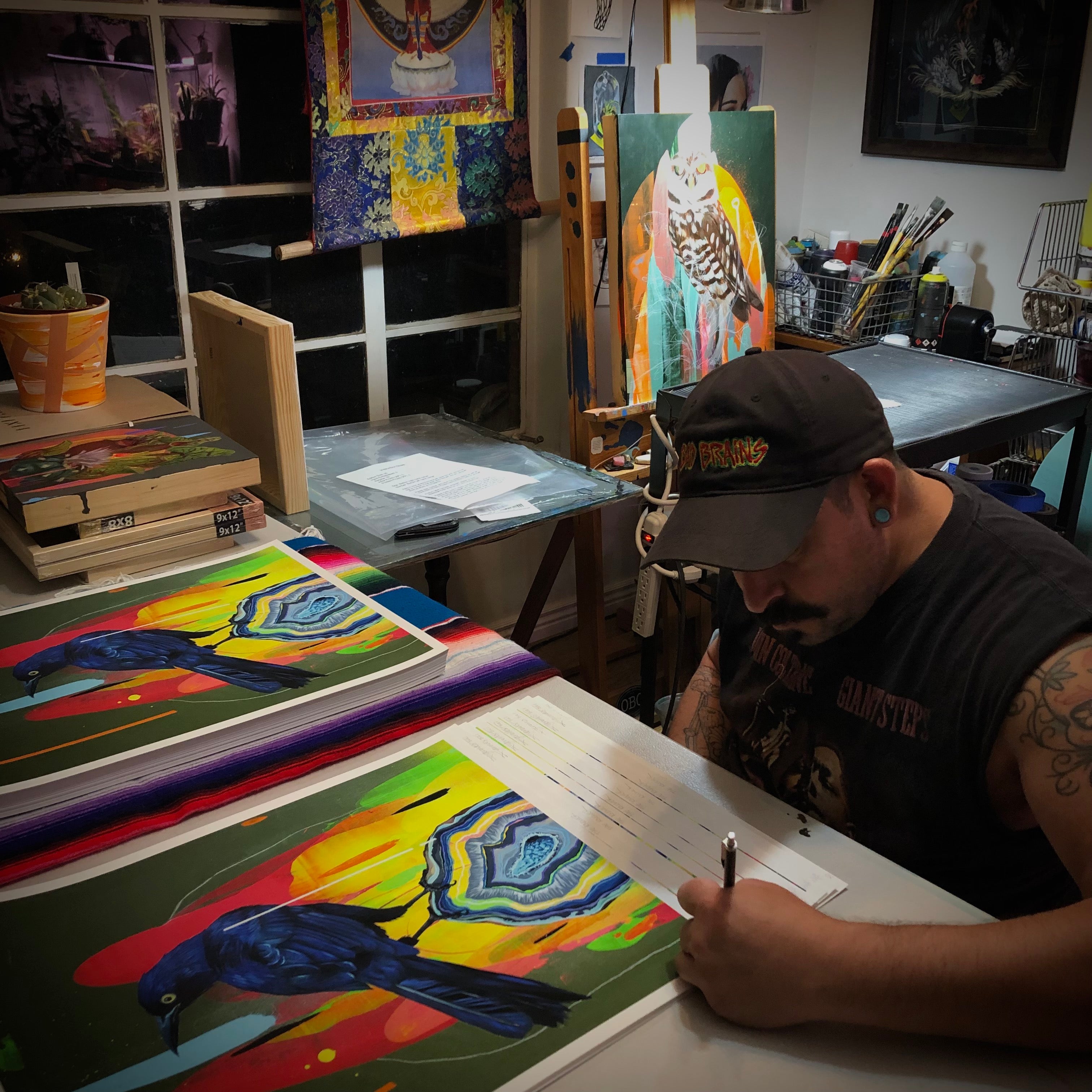A tattooed man sits at a table signing and numbering archival pigment prints of birds. The room, filled with art supplies and paintings, features a large owl piece. Warm lighting crafts an inviting atmosphere akin to Frank Gonzales The Knower studio.