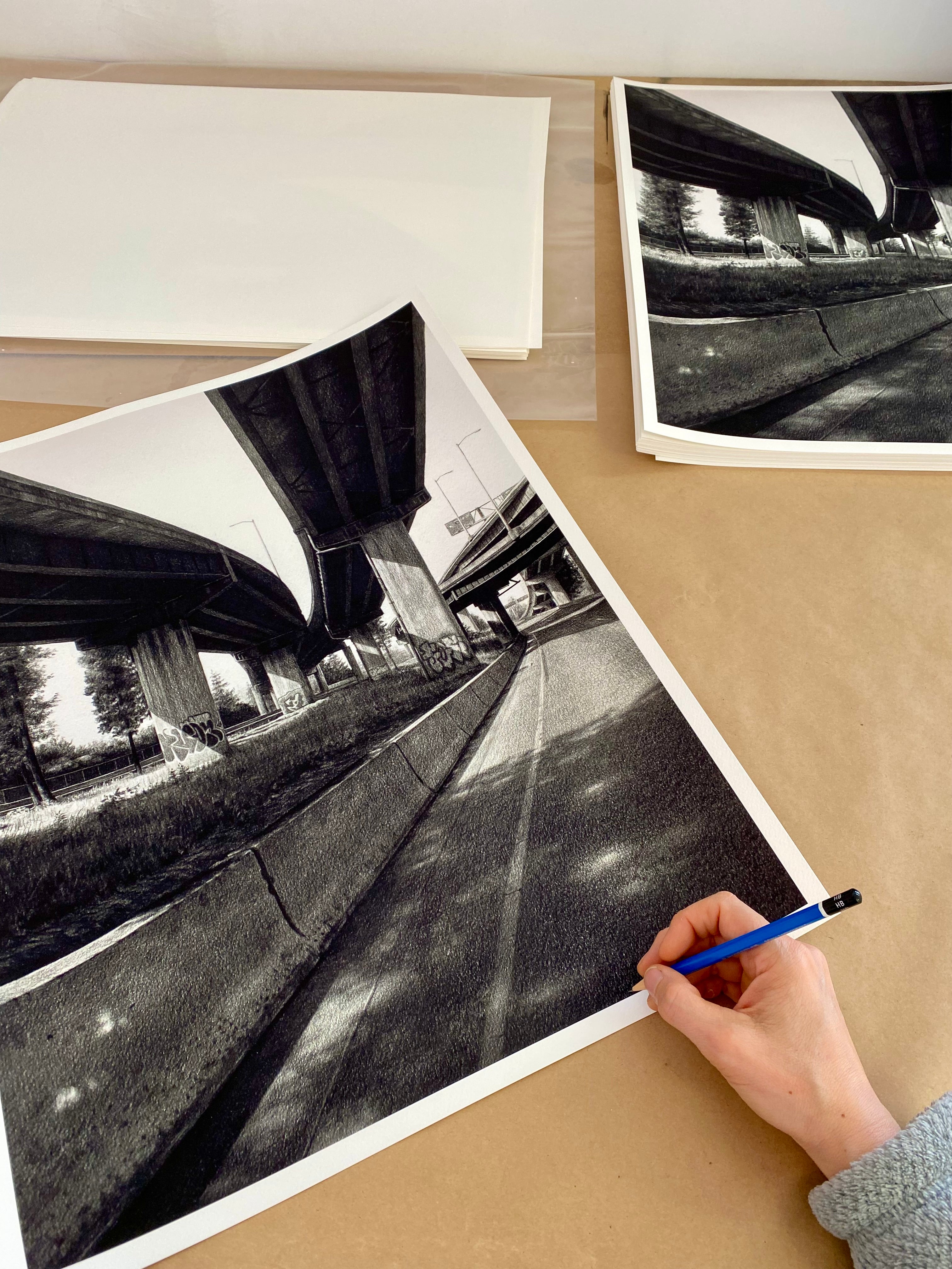 A person is sketching a detailed black and white scene of an elevated highway, complete with overpasses and grass, using a blue pen. Their work mirrors the meticulous style found in Stephanie Buers acclaimed 5 North releases.