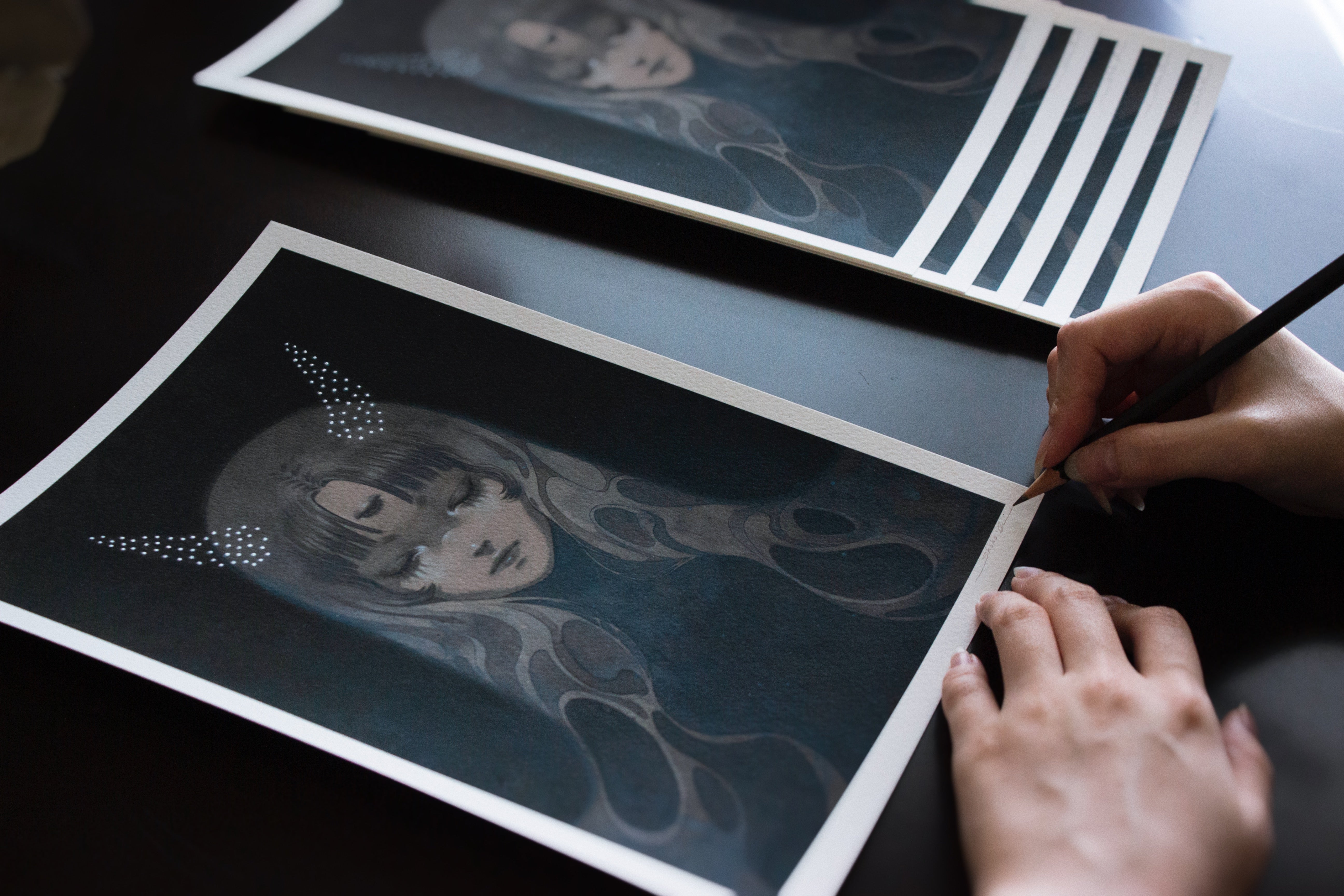 Someone is using a pencil to sign an archival pigment print by Shoko Ishida titled Stardust Slumber. The artwork shows a horned figure amidst intricate designs on a dark background. Several copies are neatly stacked on the table.