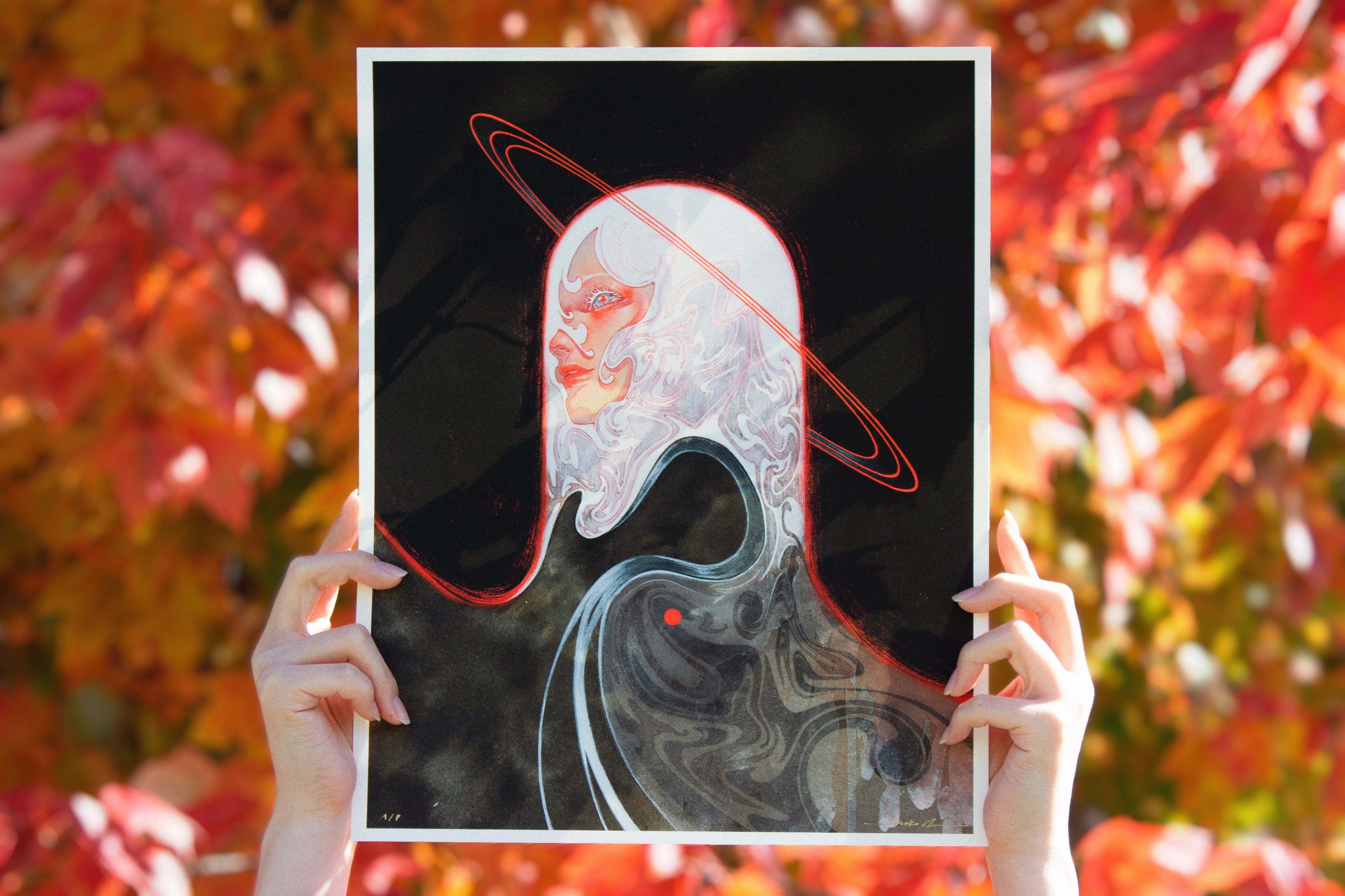 A person holds Meguri, a signed and numbered surreal art print by Shoko Ishida, showing a figure with flowing white hair and a red halo on black, set amidst vibrant orange and red autumn leaves.
