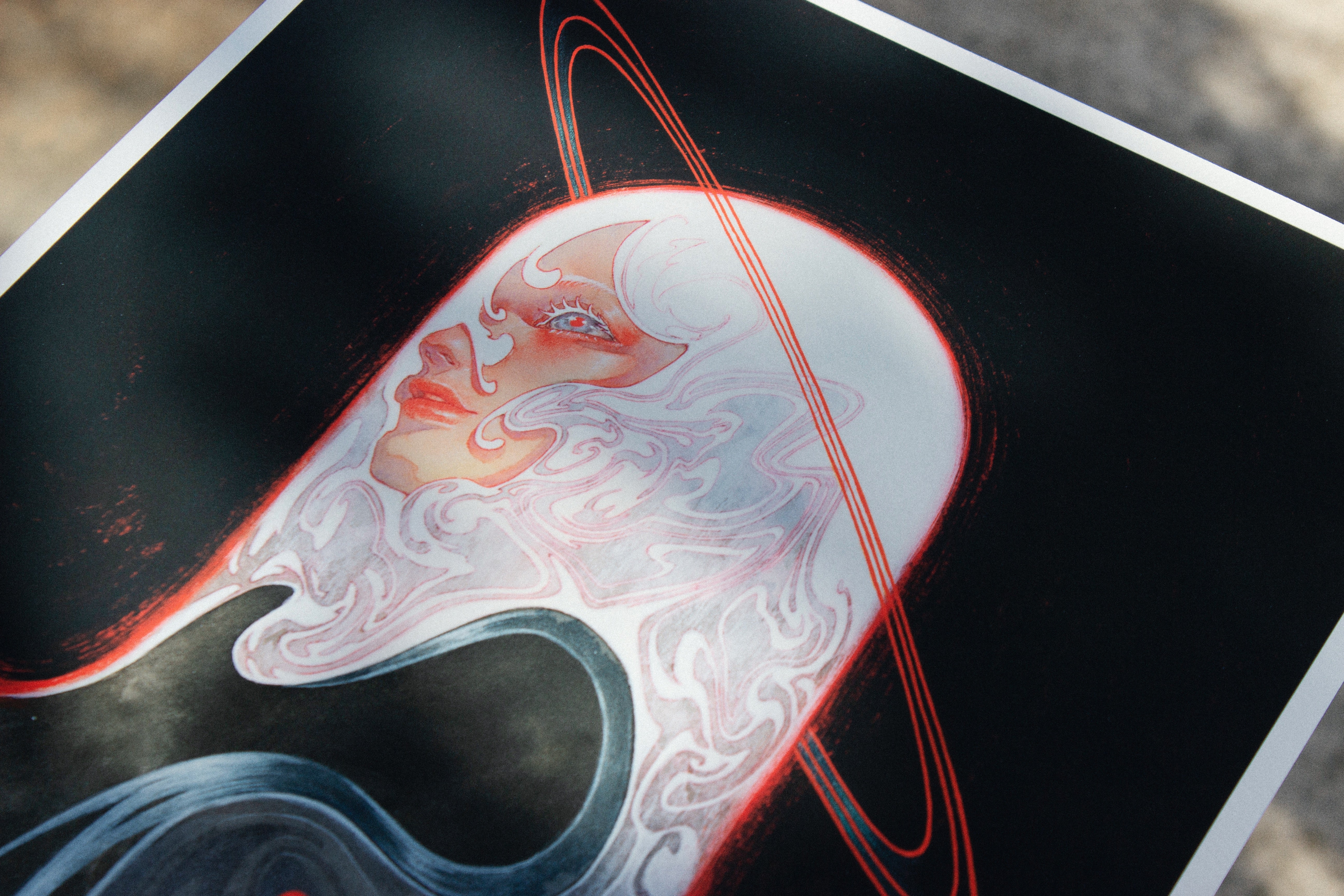 The Meguri by Shoko Ishida features a stylized figure with flowing white hair, a serene expression, and red and white accents. A red ring encircles their head against a dark background, creating a celestial or futuristic effect. This archival pigment print is signed and numbered for authenticity.