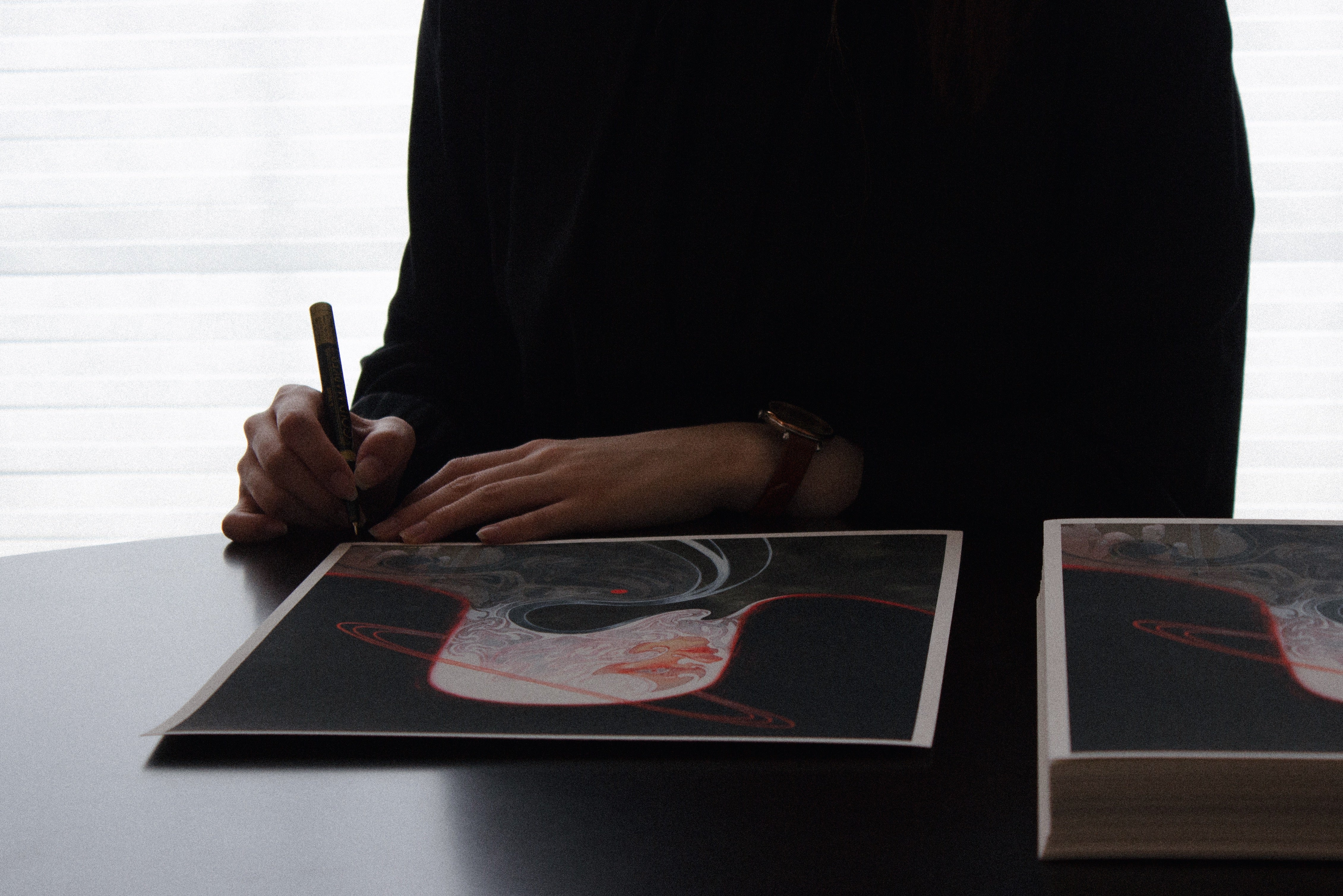 Sitting at a table, perhaps Shoko Ishida is seen holding a pen over an artwork titled Meguri from her brand. The archival pigment print features abstract red and gray swirls, is signed and numbered. A dark long-sleeve shirt is partly visible while blinds cover the tall window behind.