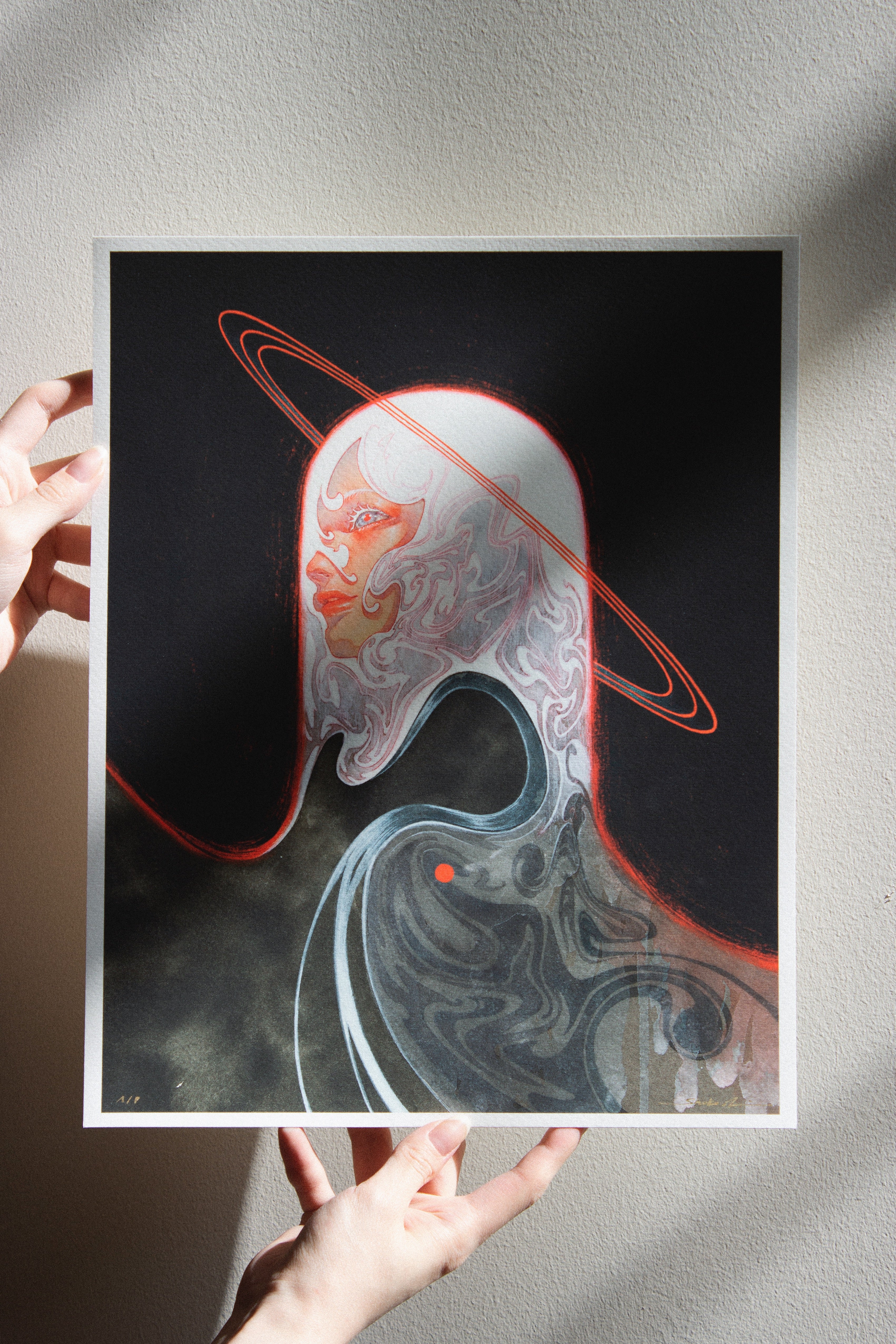 A person holds Meguri, an archival pigment print by Shoko Ishida, featuring a surreal figure with long white hair on a dark background. A red halo encircles the head amidst abstract shapes, all signed and numbered for authenticity.