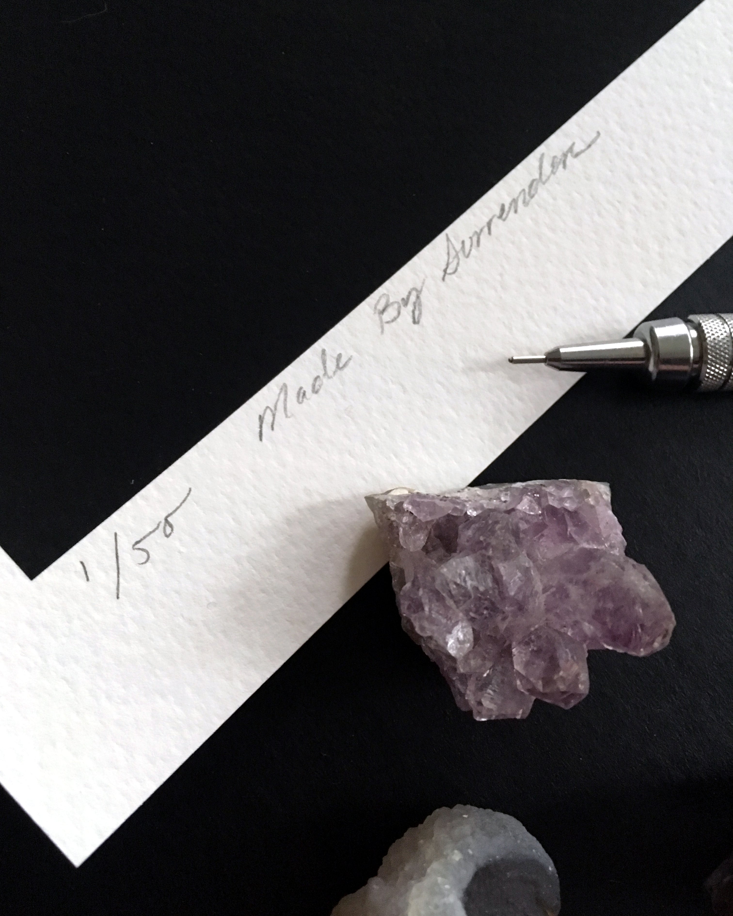 A textured white paper on black surface displays handwritten Made By Surrender and 1/50, set near a purple crystal and mechanical pencil, hinting at an Archival Pigment Print by Christina Mrozik.