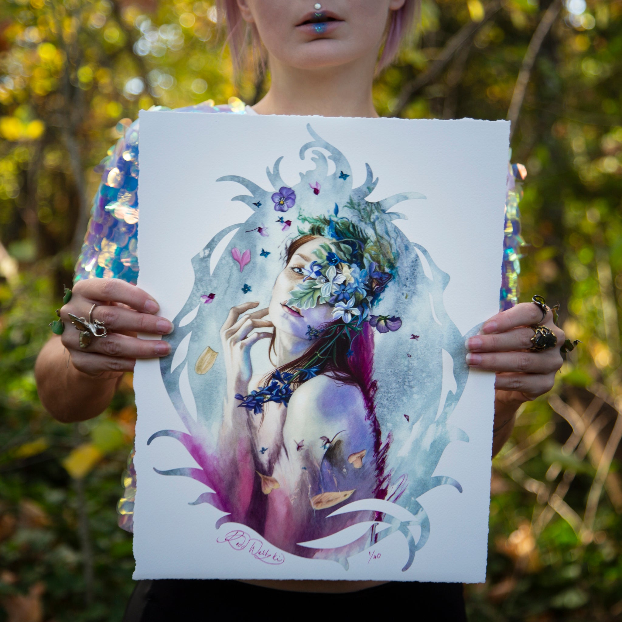 A person holds Veiled in Springtime by Redd Walitzki, showcasing a woman with floral adornments against a blurred forest backdrop. The limited edition print features vivid colors and intricate details that partially conceal the holder’s face behind its vibrant beauty.