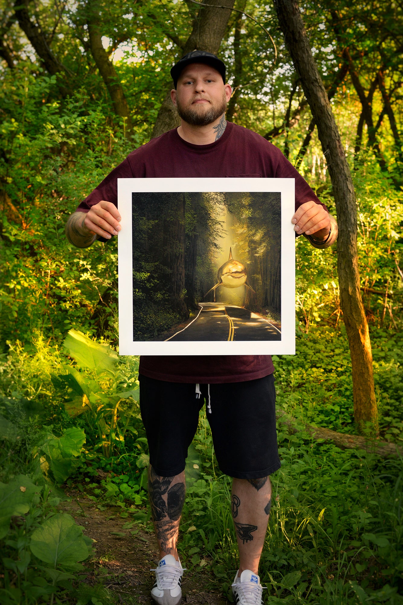 In a sunlit forest, a man in dark attire holds Rewarded Are The Curious, an archival pigment print by Chris Austin featuring a cat in a wizard hat with a forest backdrop, surrounded by lush green foliage.