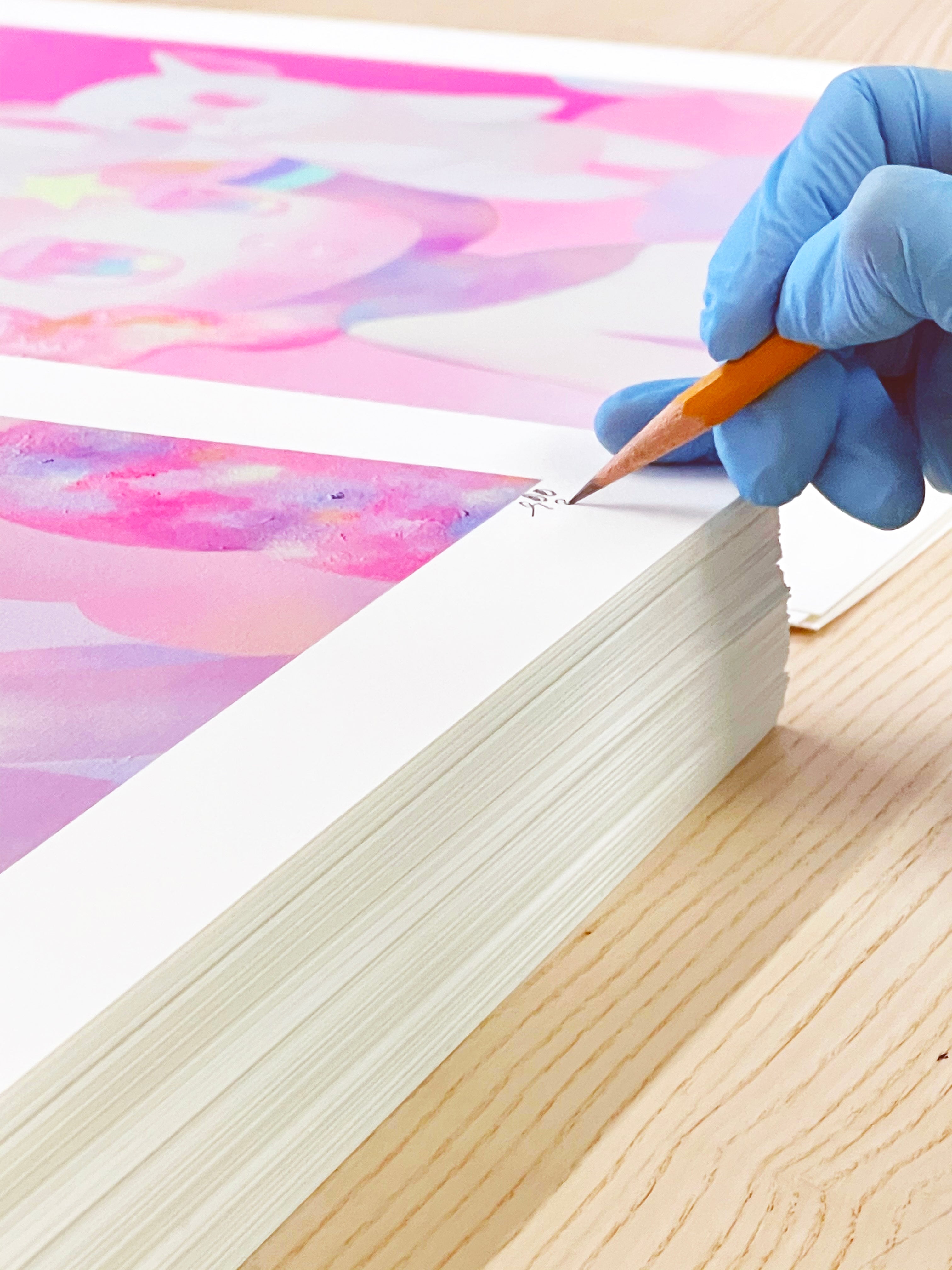 Wearing blue gloves, a person signs a stack of the limited edition Hide & Seek archival pigment prints by So Youn Lee with a pencil on a light wooden table, bringing the vibrant pink and purple hues to life.