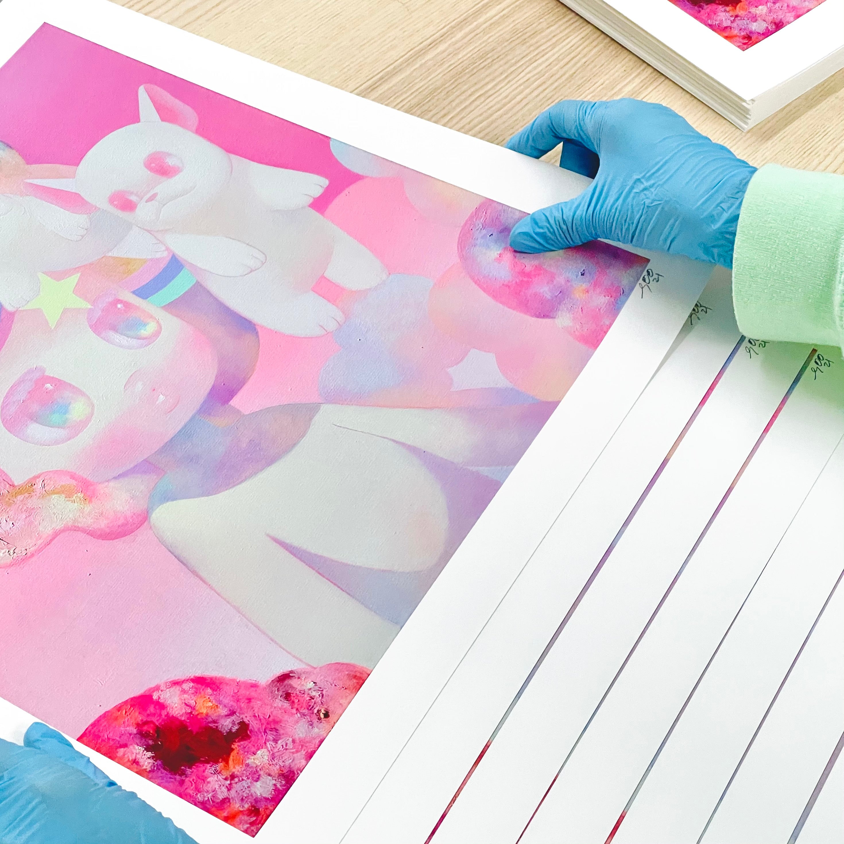 Wearing blue gloves, someone carefully handles Hide & Seek, a So Youn Lee limited edition pastel art print featuring a whimsical creature with large eyes and a small winged companion against a soft pink cloud background. Archival pigment prints are stacked on wooden surface.