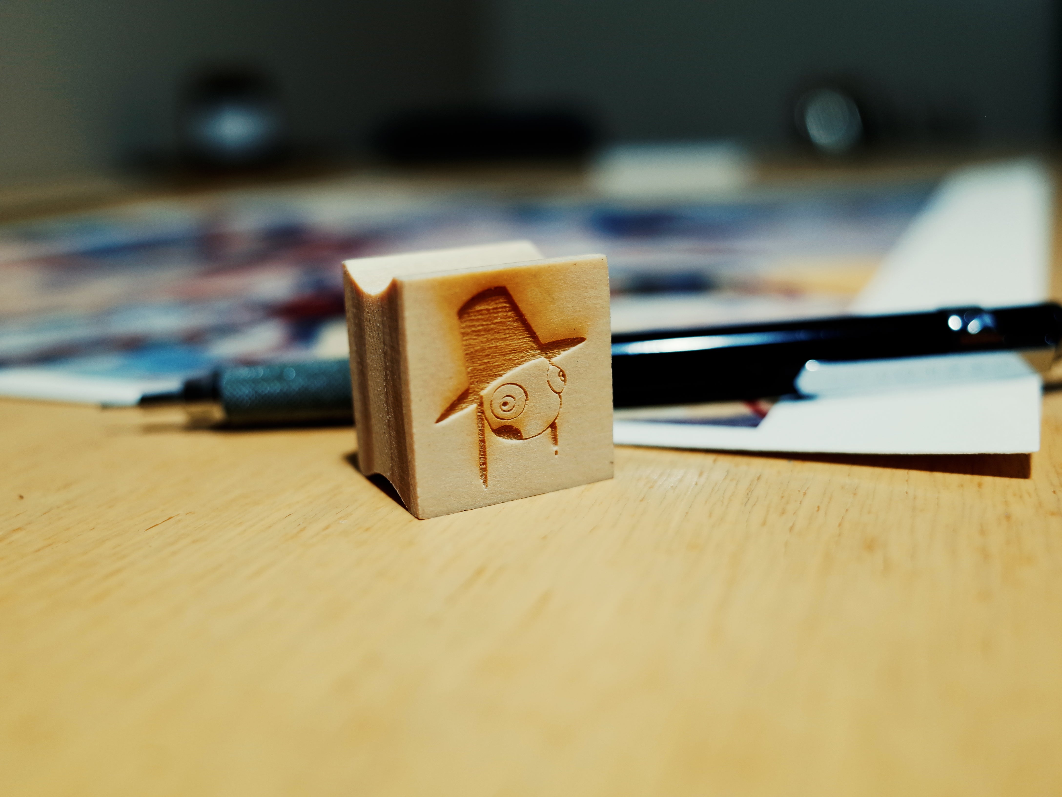 A wooden stamp titled Festival (마당놀이) by Young-Ji Cha, showcasing a playful face with sunglasses and a hat, sits on the table. Nearby, a pen and softly blurred papers enhance the warm ambiance reminiscent of Young-Ji Chas artistic style.